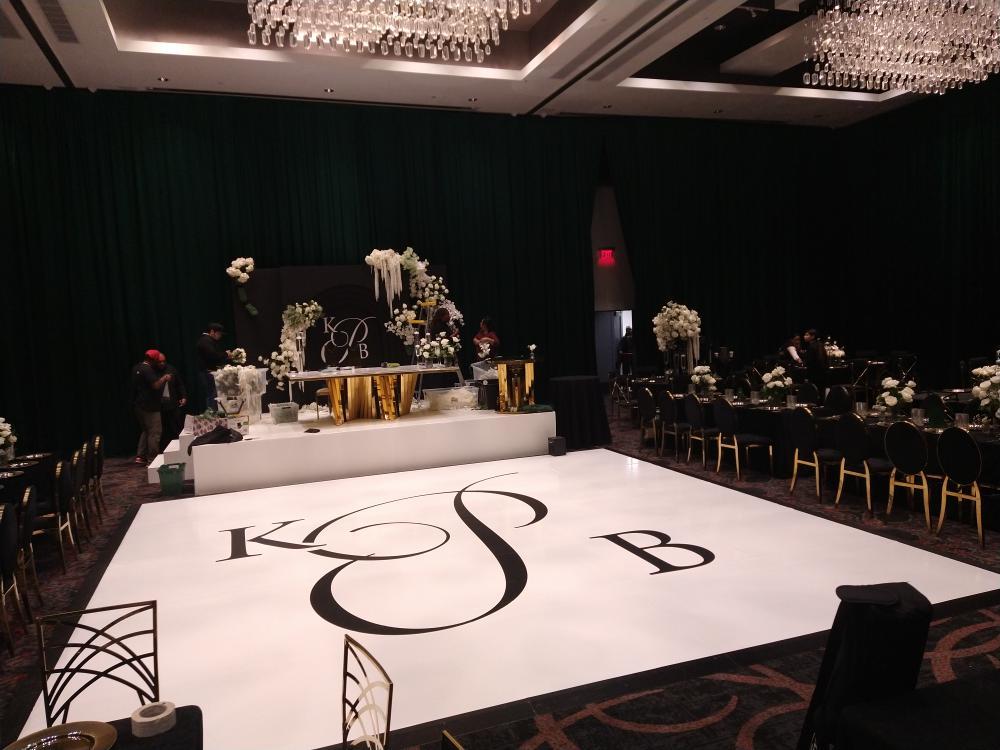 Event space with a decorated table, floral arrangements, and a large white dance floor with initials, surrounded by tables and chairs.