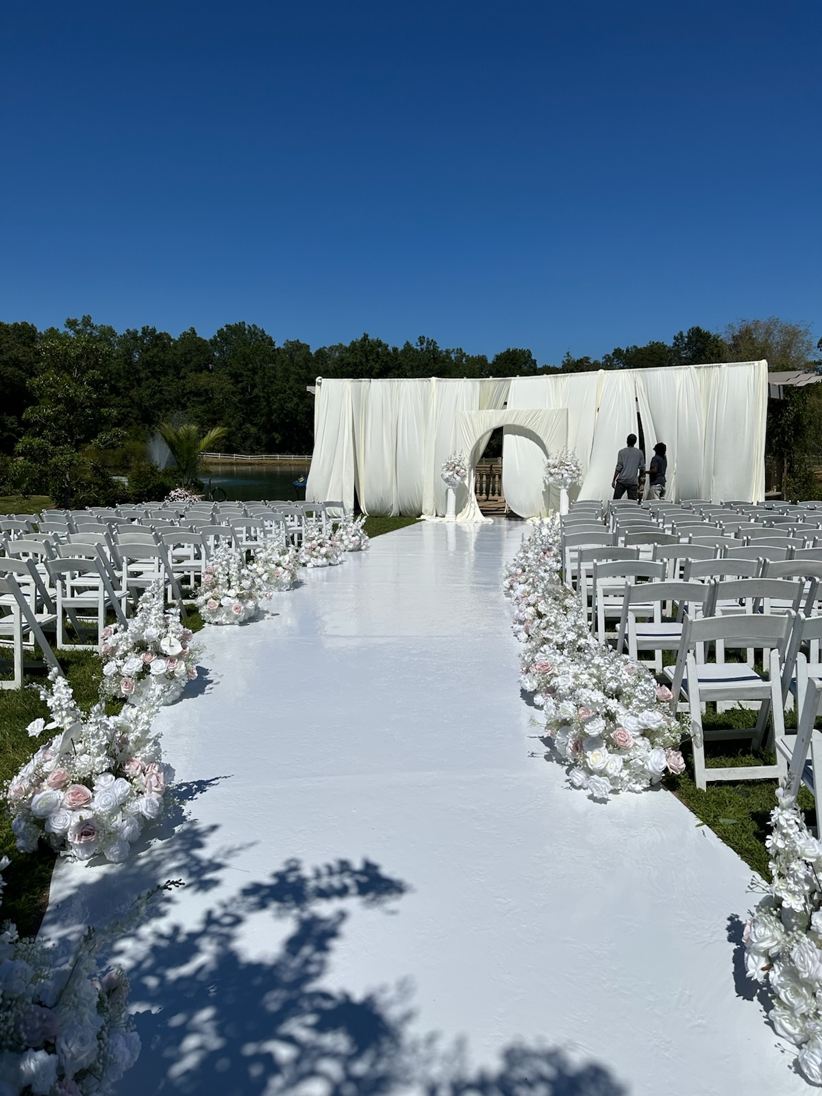 Outdoor wedding setup with white chairs, floral decorations, and a white aisle leading to a circular arch against a blue sky.