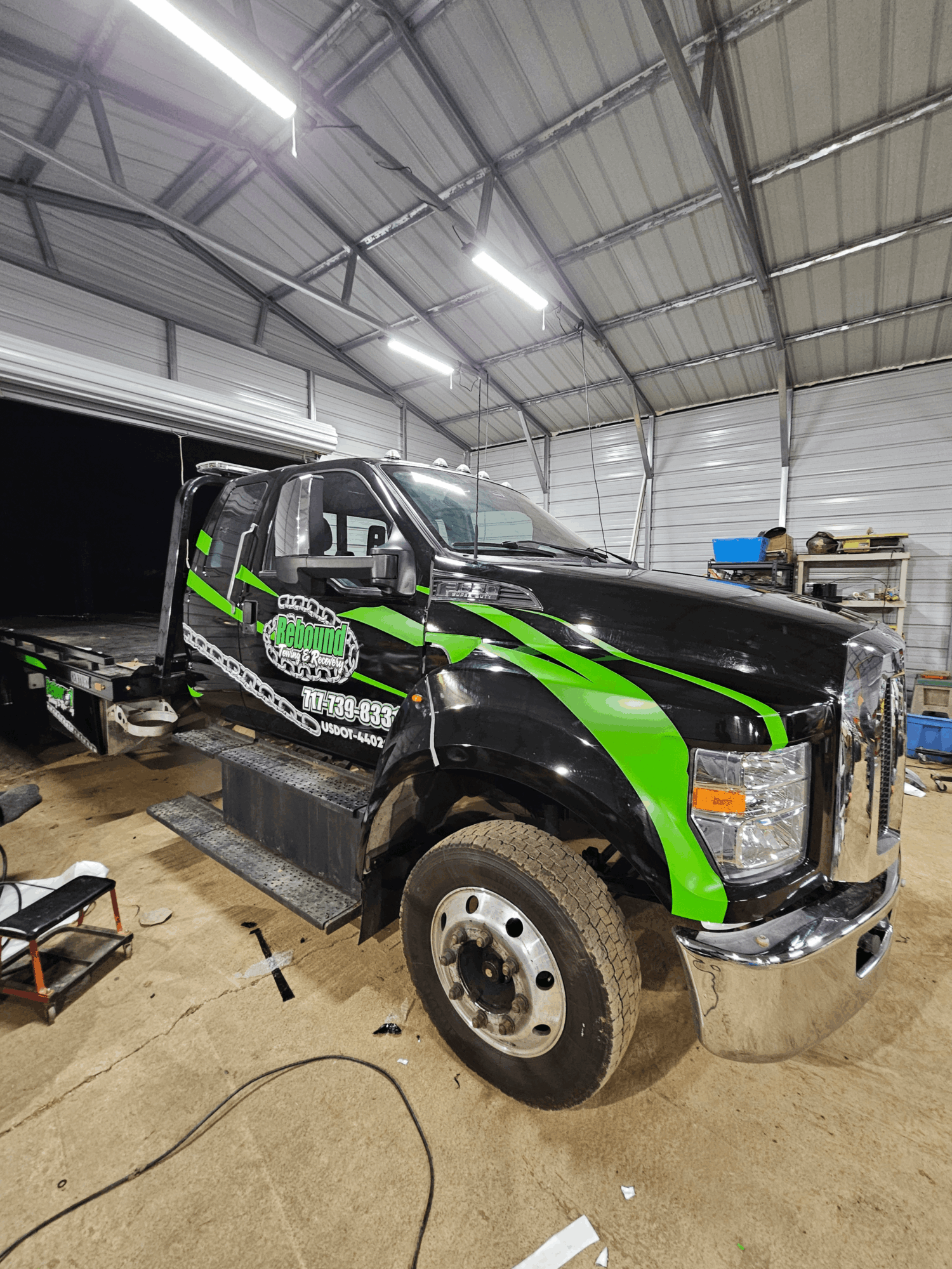 Black and green semi-truck inside a warehouse with a curved metal roof, fluorescent lighting, and various tools and equipment around.