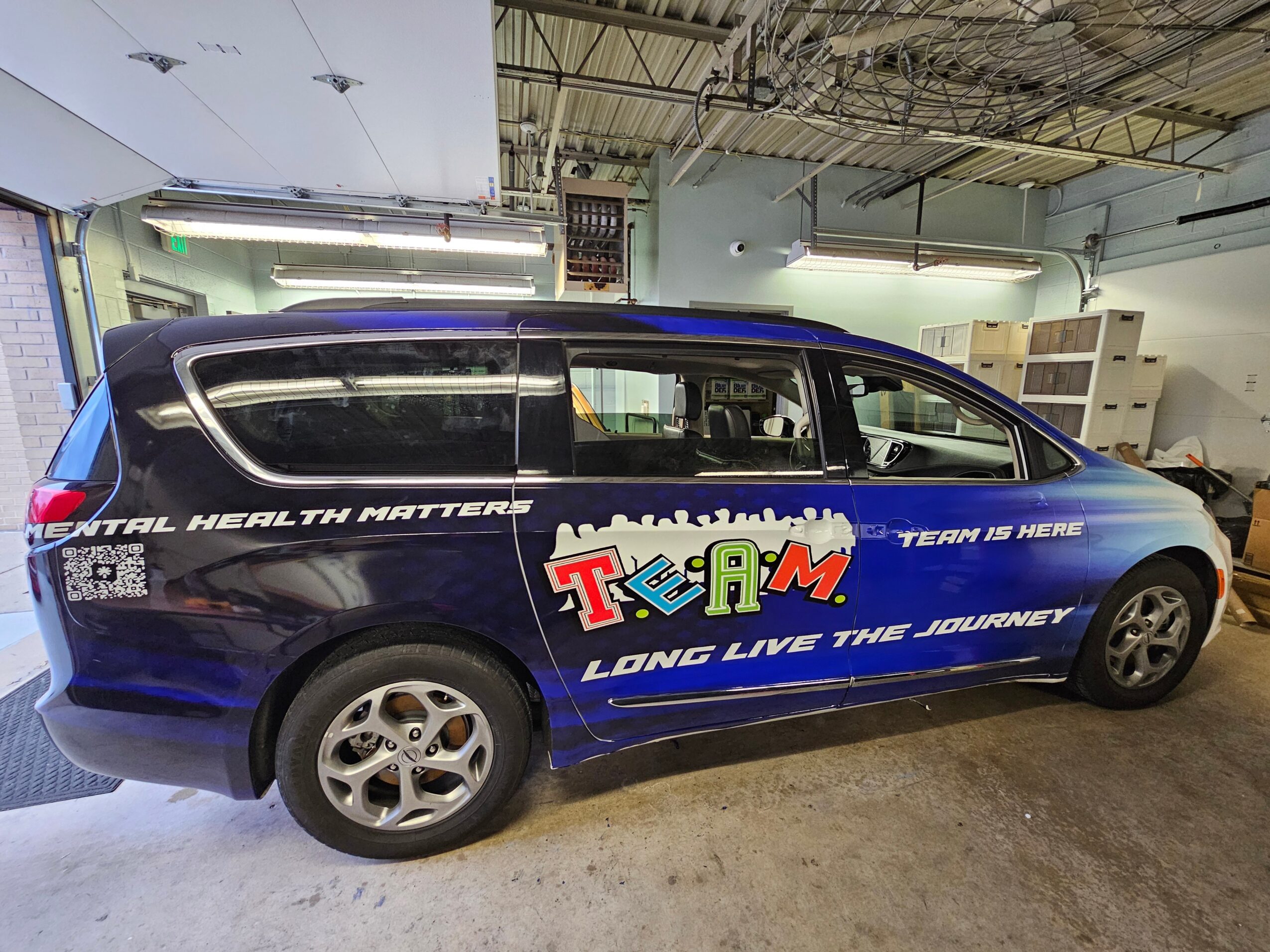 Blue van with colorful TV and team graphics, parked indoors with ceiling and storage shelves visible.