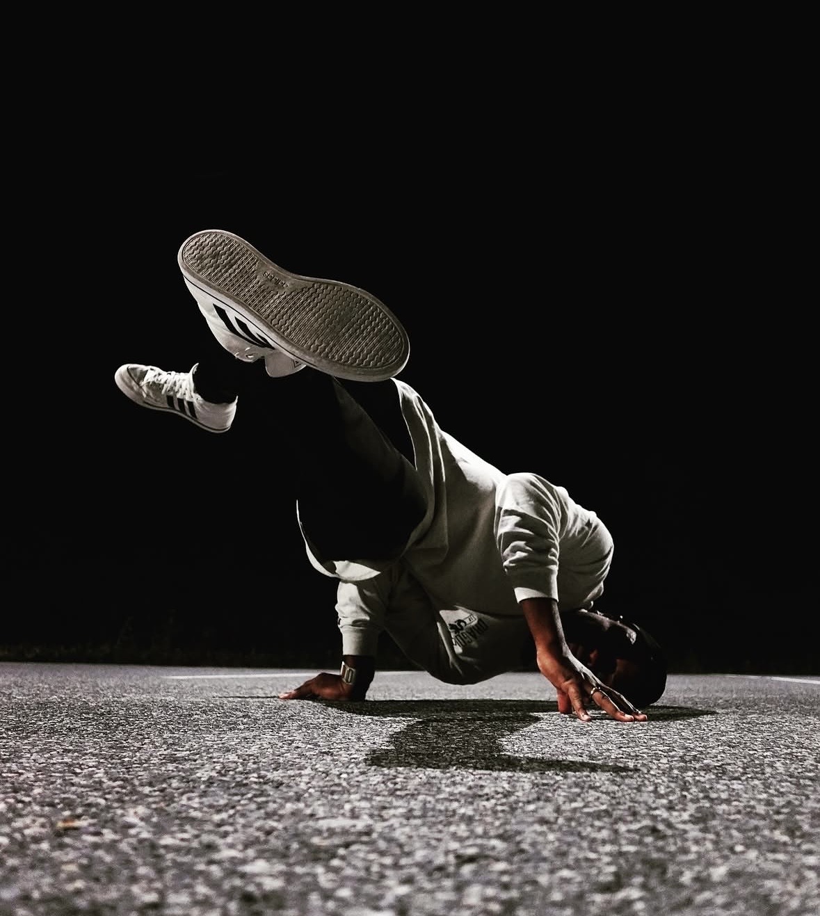 Person performing a breakdancing move on the ground with one hand and head, legs raised in the air, against a black background.