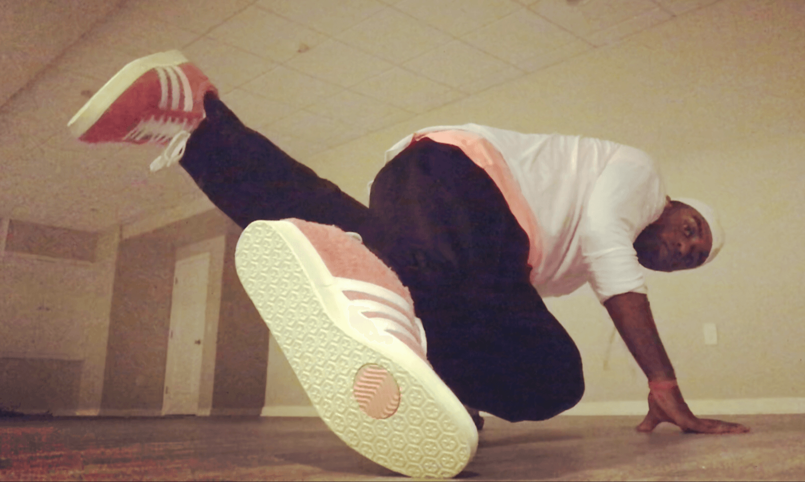 Person in a white shirt and black pants crouches on the floor with one leg extended upward, wearing pink sneakers.