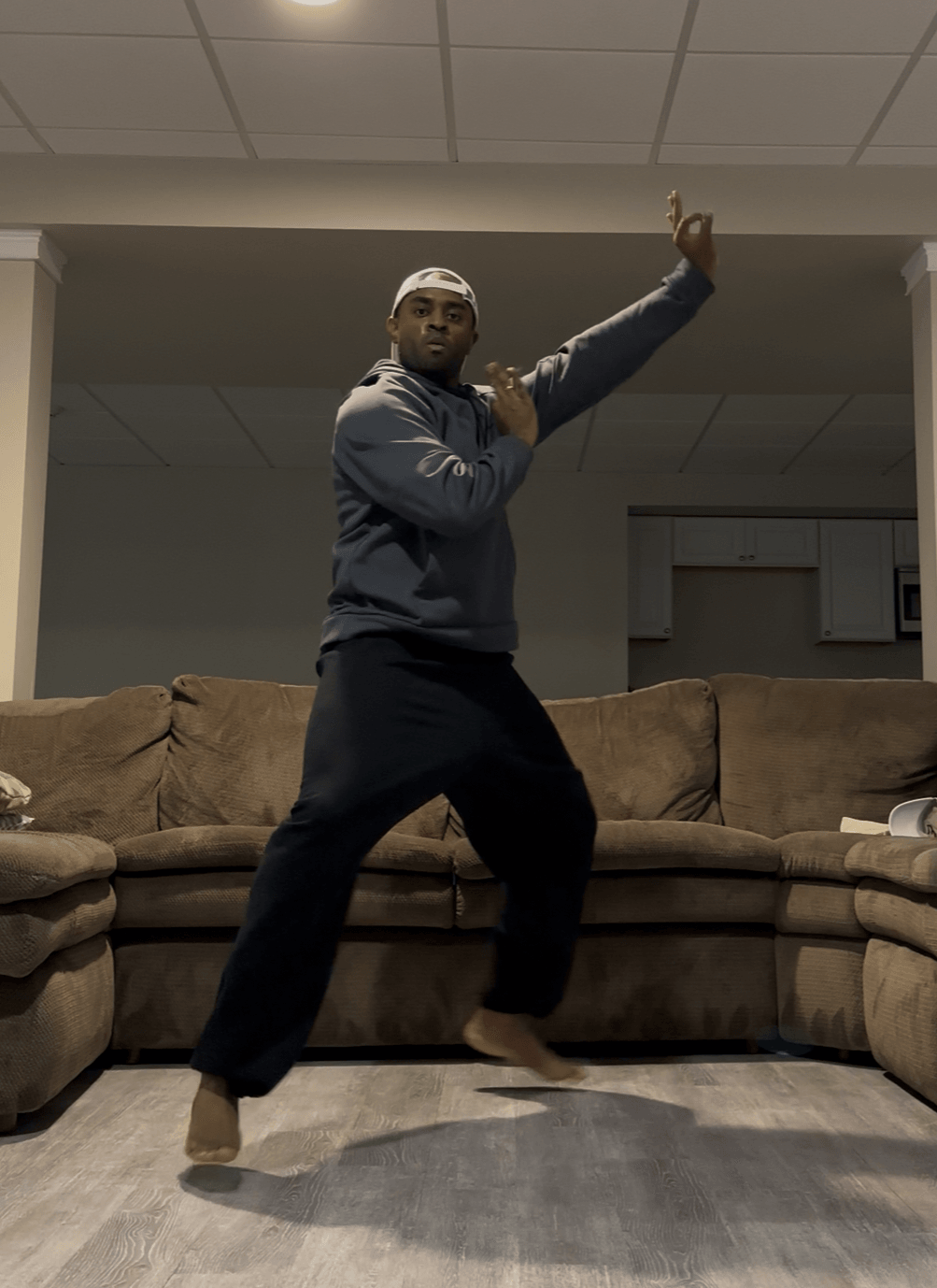 Person standing on a couch with one arm raised and the other hand on chest, in a room with ceiling tiles.
