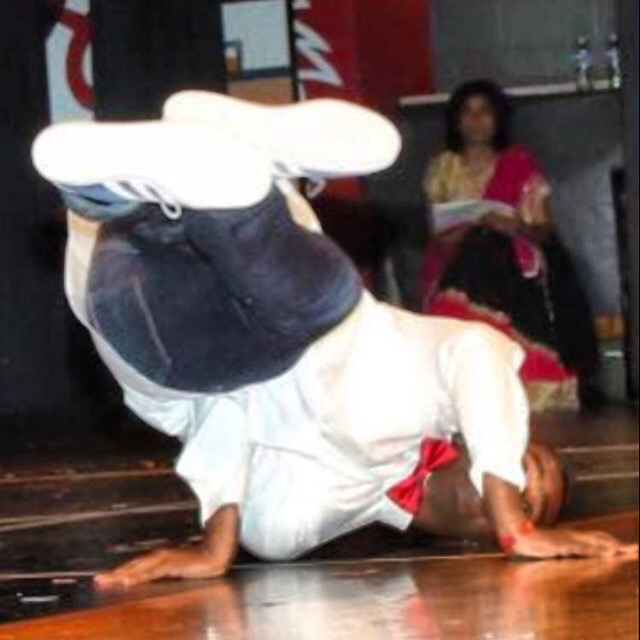 Child performing a headstand on the floor with a person sitting in the background holding a book.