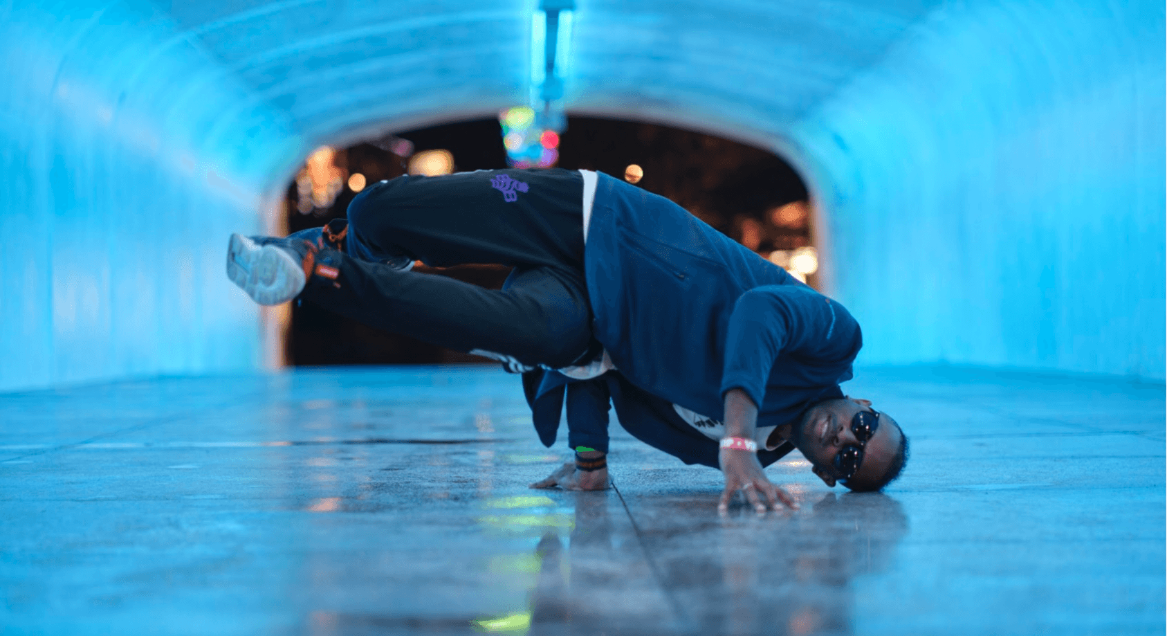 Person in dark clothing performing a headstand in an illuminated tunnel at night, with city lights in the background.
