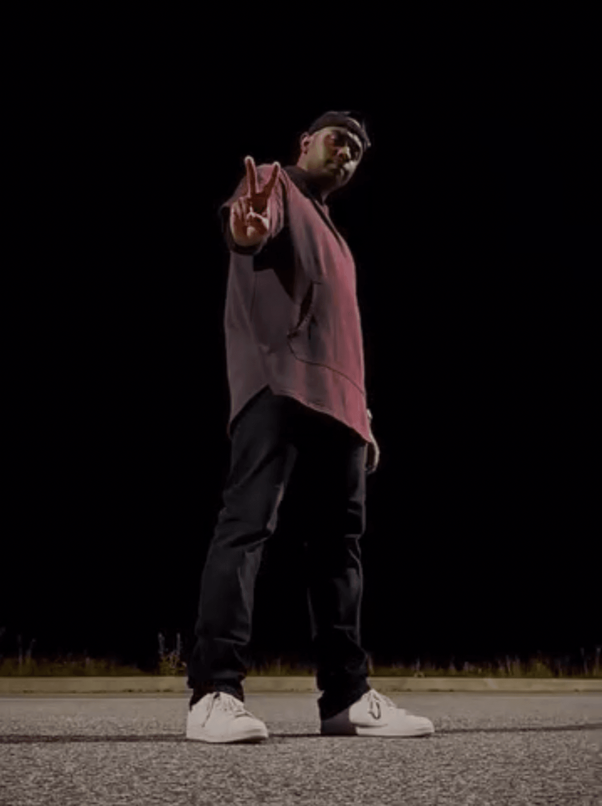 Person standing on road at night, pointing forward, wearing a hoodie and sneakers, with dark background.