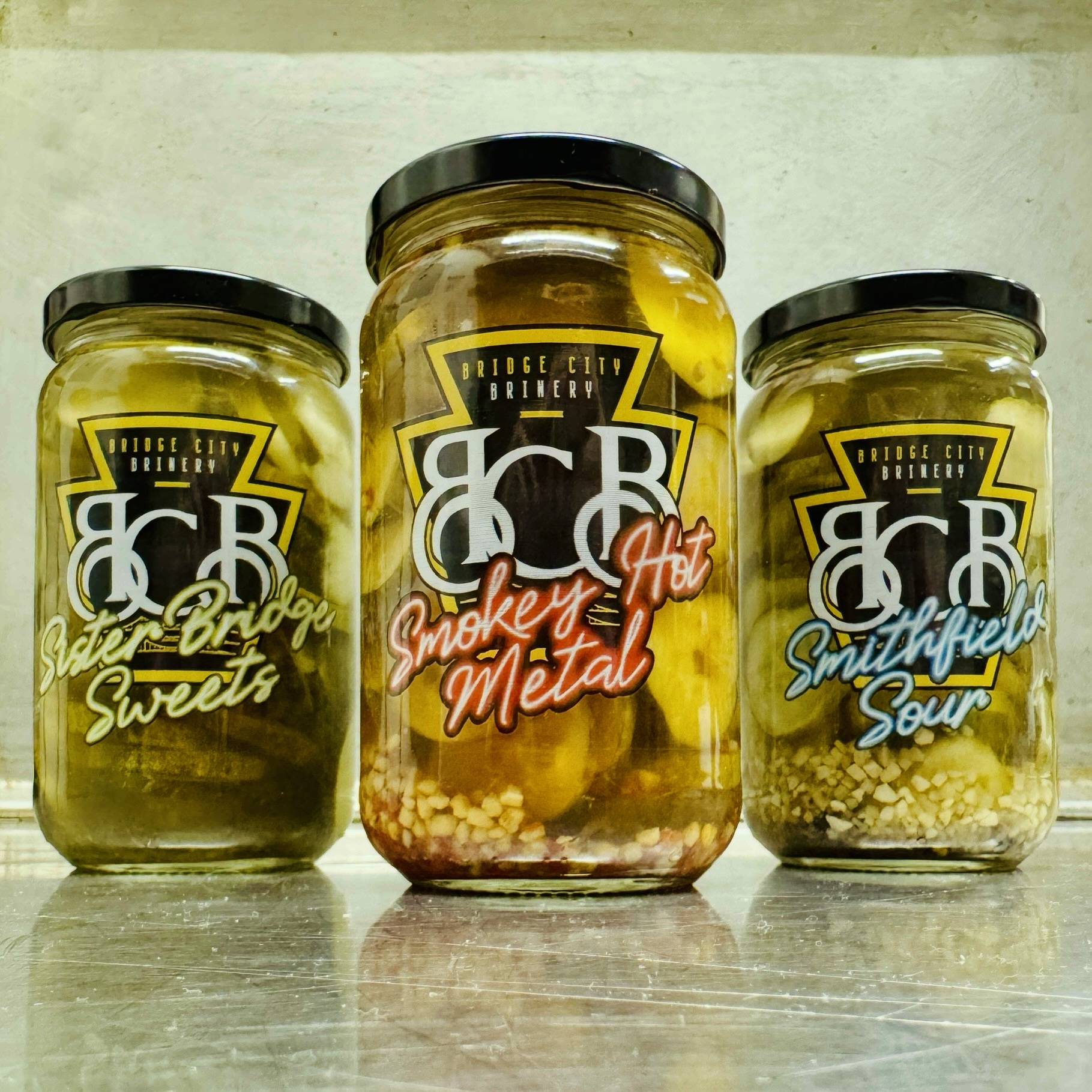 Three jars with black lids, labeled as sweets, smoky hot metal, and sour, placed on a surface against a plain background.