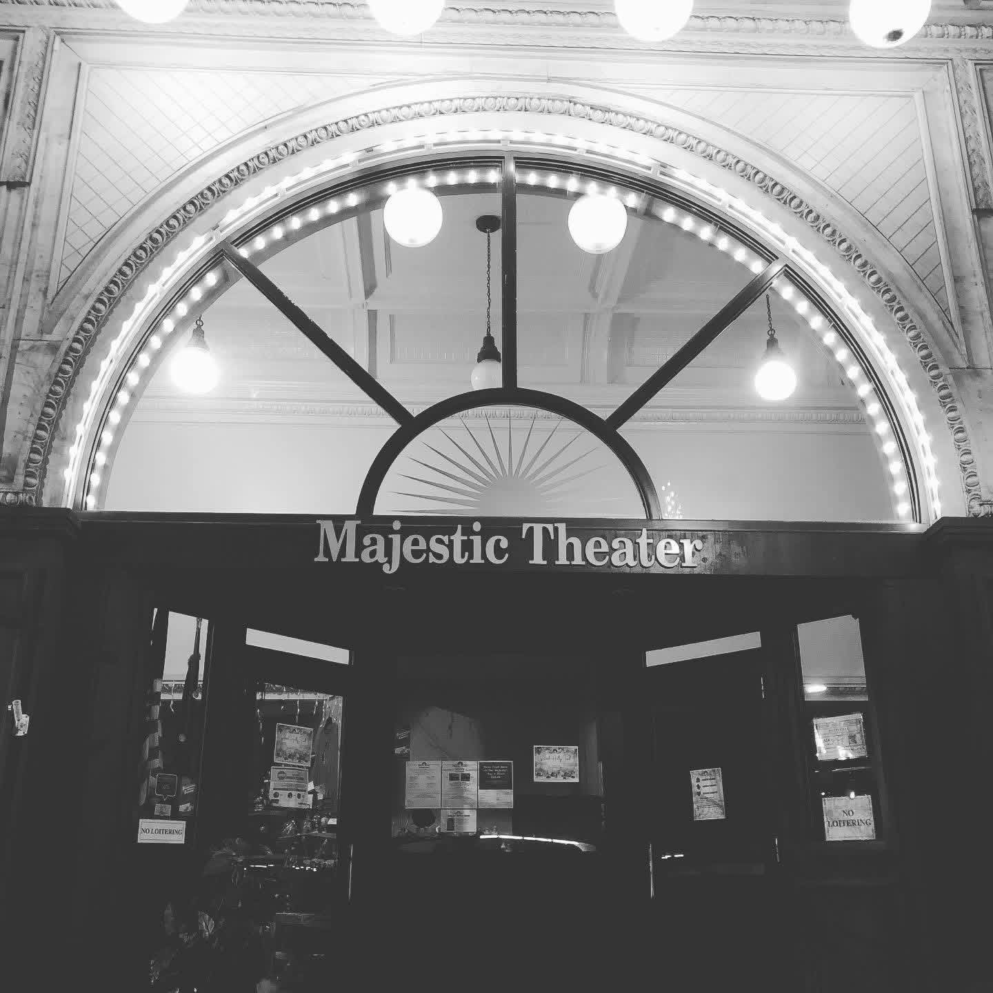 The entrance of Majestic Theater with a large clock above the door, illuminated by hanging lights, in black and white.