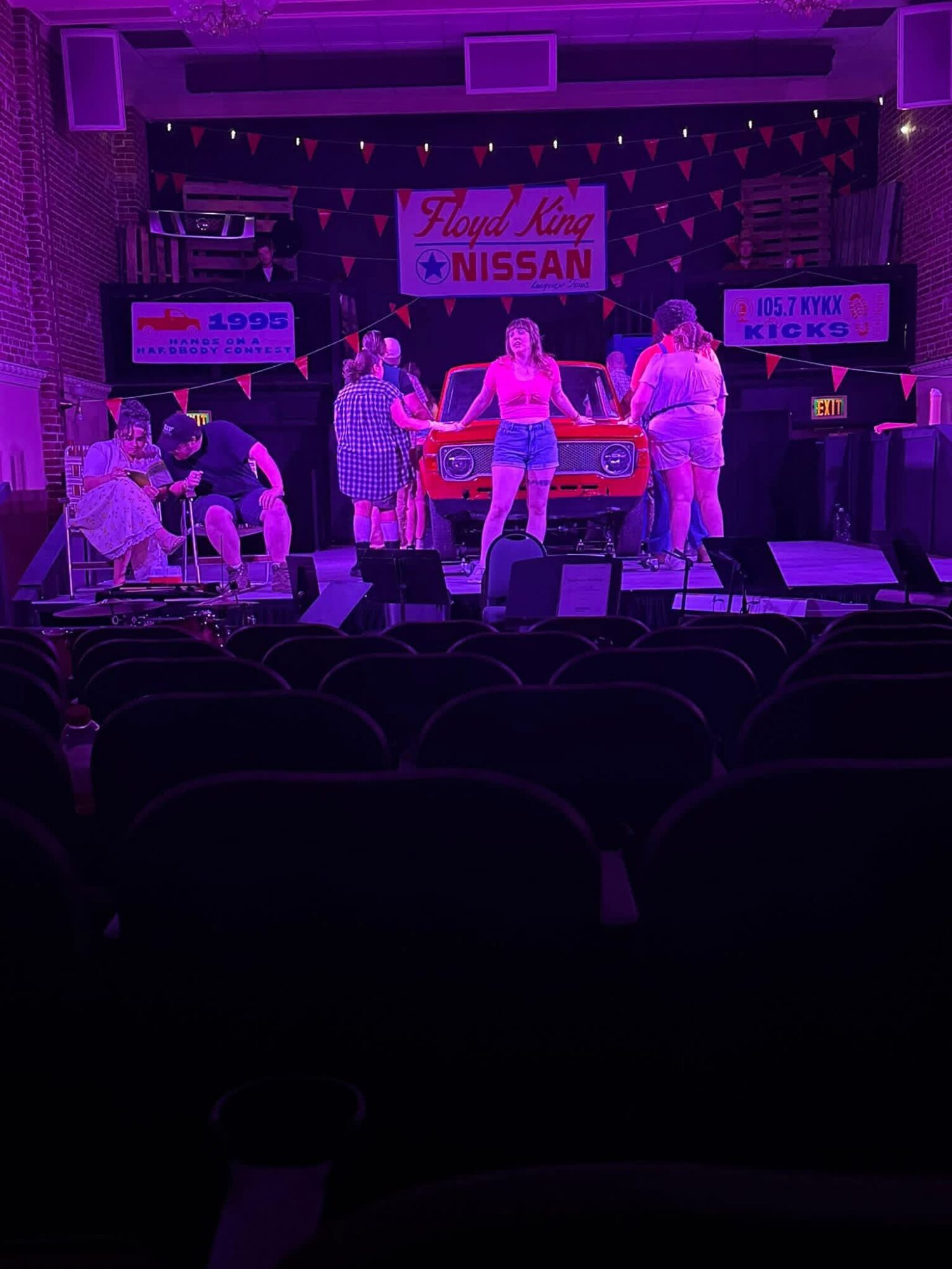 Stage with people, banners, and a pool table, illuminated by purple lighting, in a theater or event space.