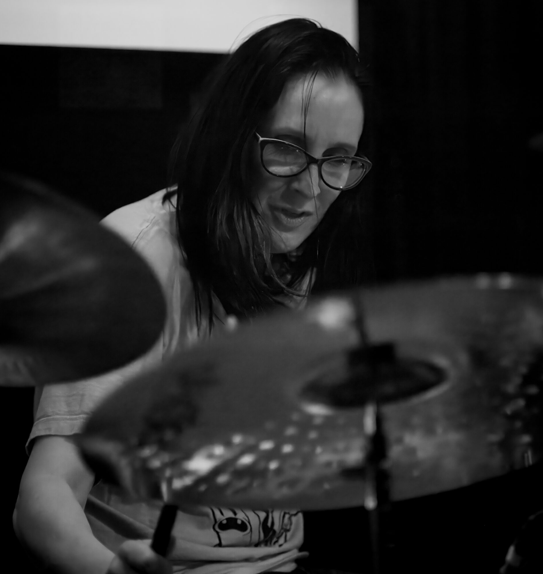 Woman with glasses playing drums, focused on cymbals, in black and white.