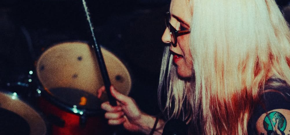 Person with long blonde hair and glasses holding drumsticks near drums, side view, dark background.
