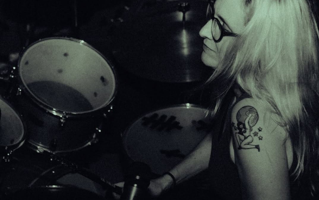 Woman with glasses and a tattoo on her arm playing drums in a dark setting.