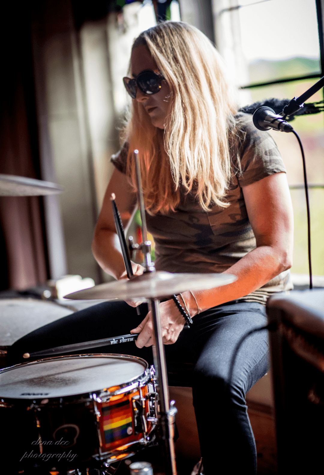 Woman with long blonde hair wearing sunglasses playing drums indoors, window with outdoor view in background.