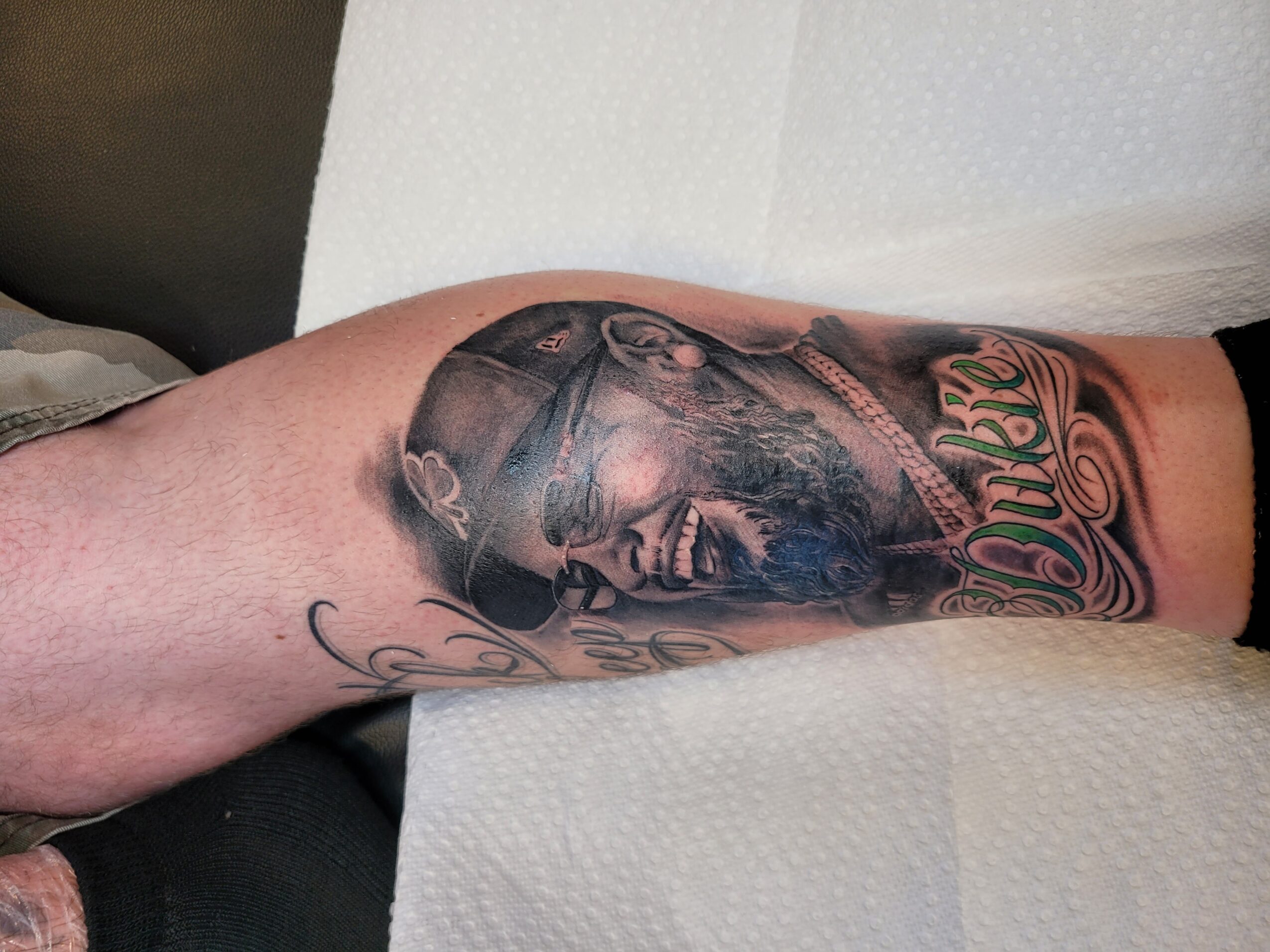 Tattoo of a man's face with sunglasses and beard on forearm, with decorative script below, on light skin.