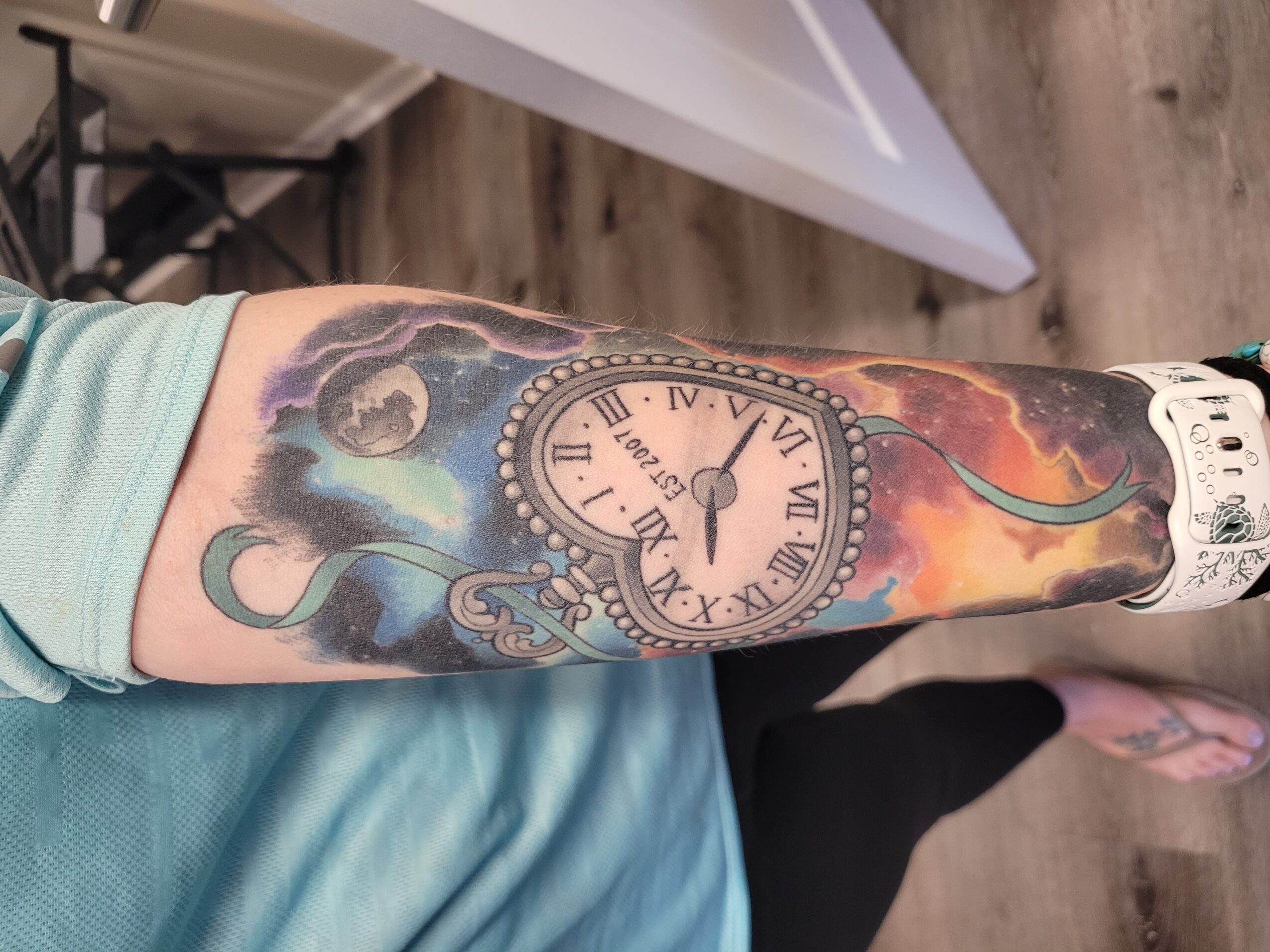 Forearm tattoo of a clock with Roman numerals, surrounded by clouds and colorful background, with a wristwatch on the wrist.