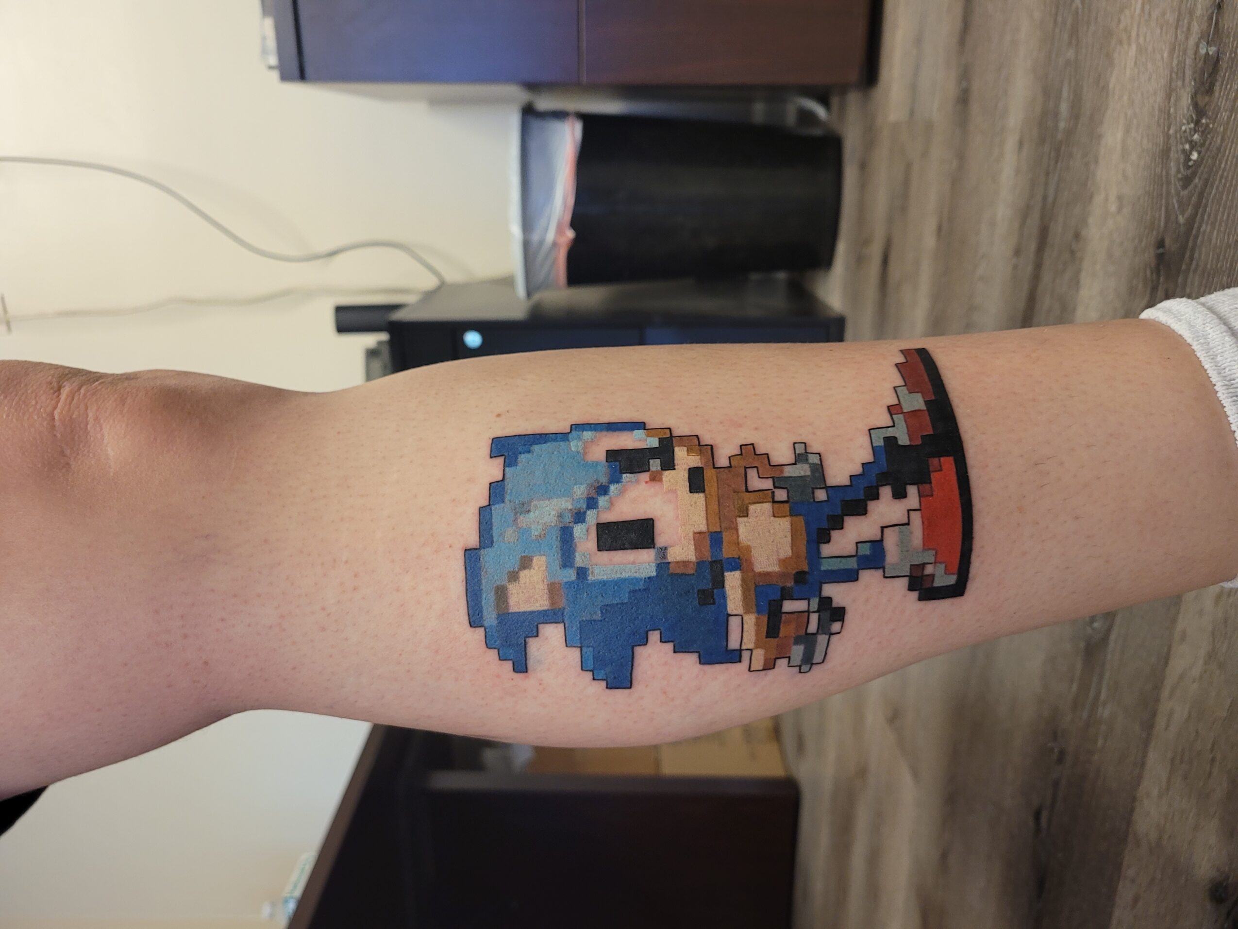 Pixel art tattoo of Sonic the Hedgehog on person's arm, with blue fur, red shoes, and a confident pose.