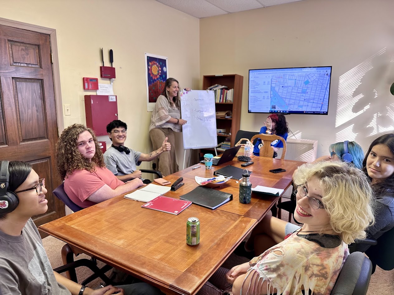 Group of people in a meeting room with a woman presenting at a whiteboard, a large screen, and a wooden table.