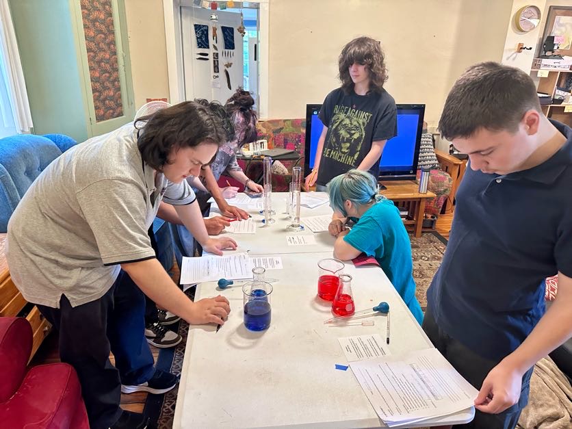 Group of children and an adult gathered around a table with jars and papers, engaging in a hands-on activity in a room.