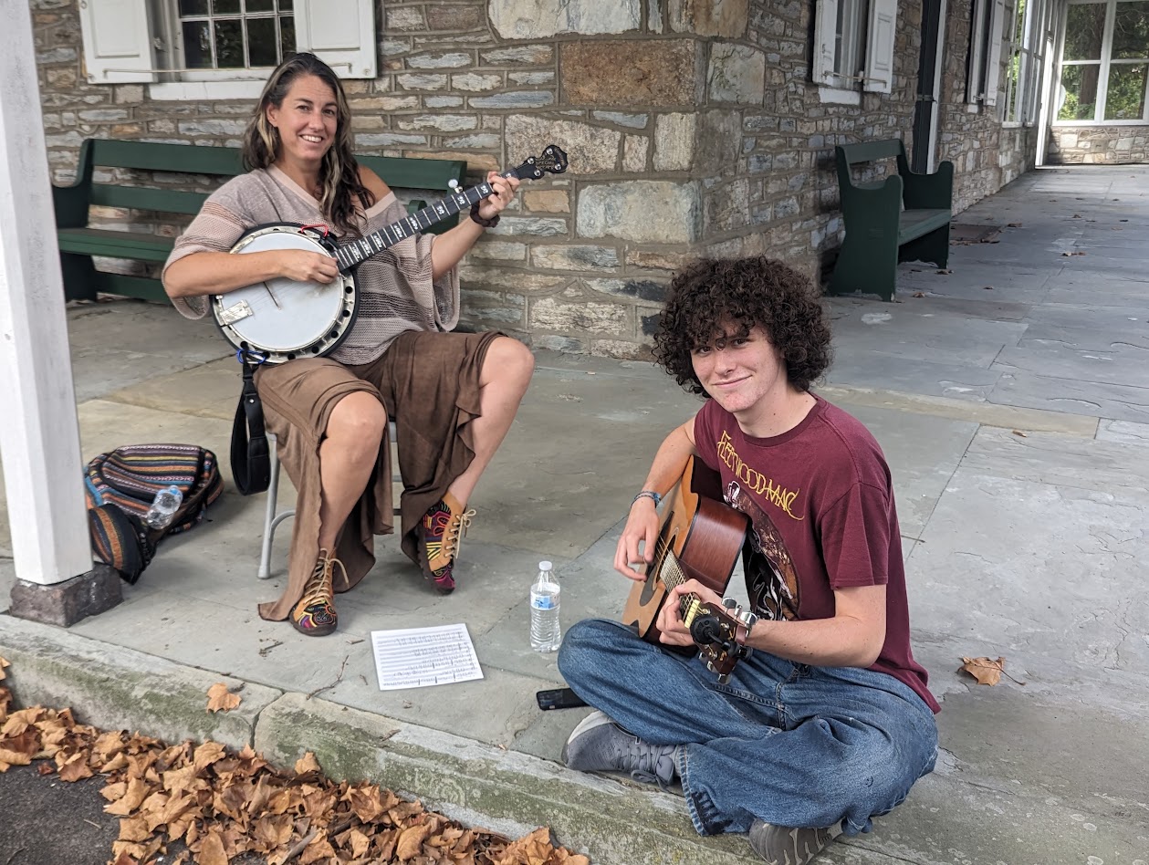 Two people playing guitars outdoors near a stone building, sitting on the ground with leaves nearby.