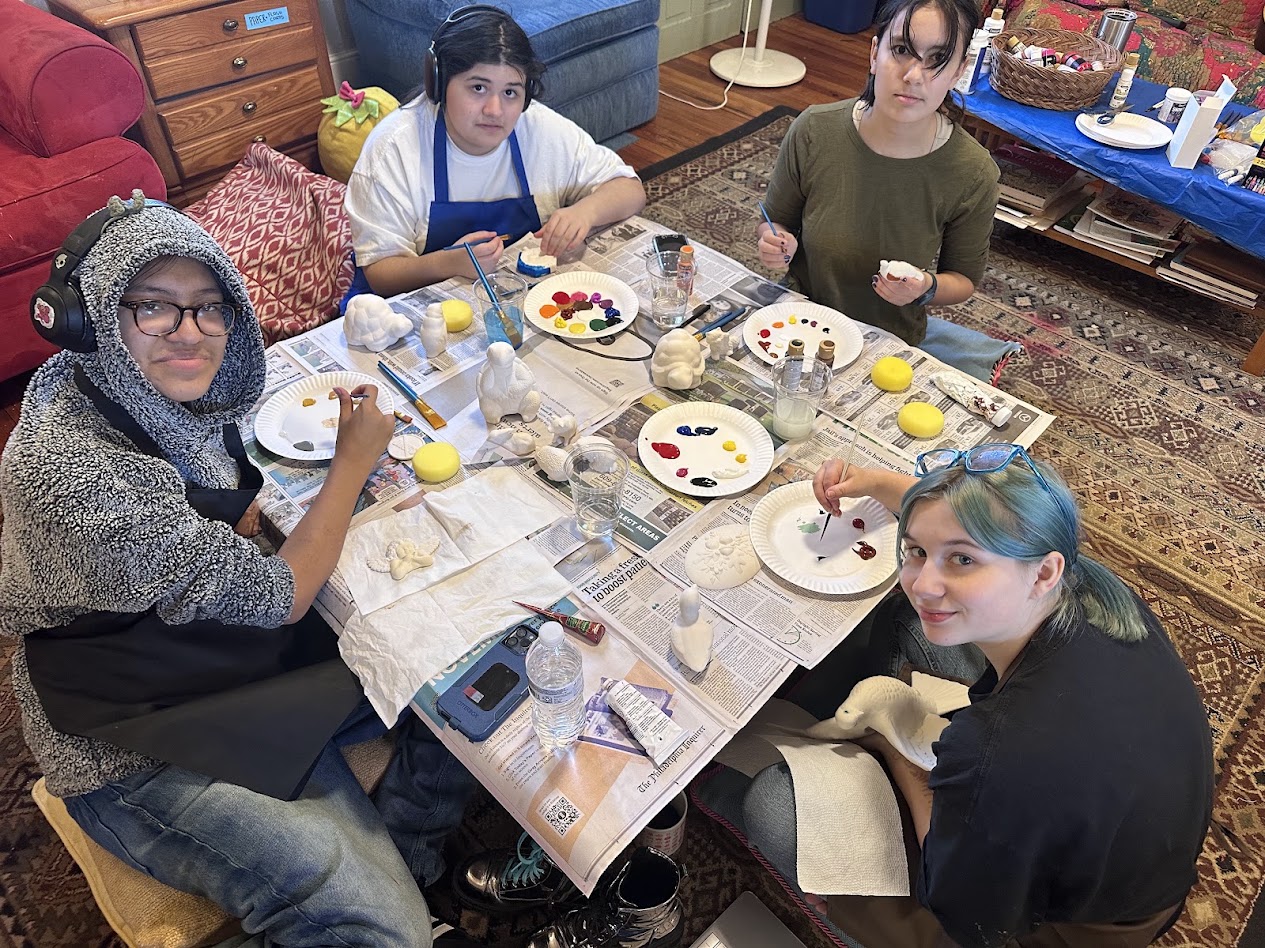Four people sit around a table painting eggs with various colors and brushes, in a cozy room with furniture and a rug.