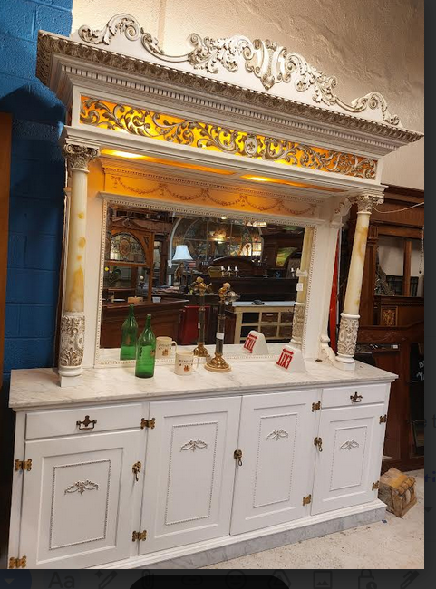White ornate mirror with decorative carvings, above a white cabinet with bottles and small objects, in a room setting.