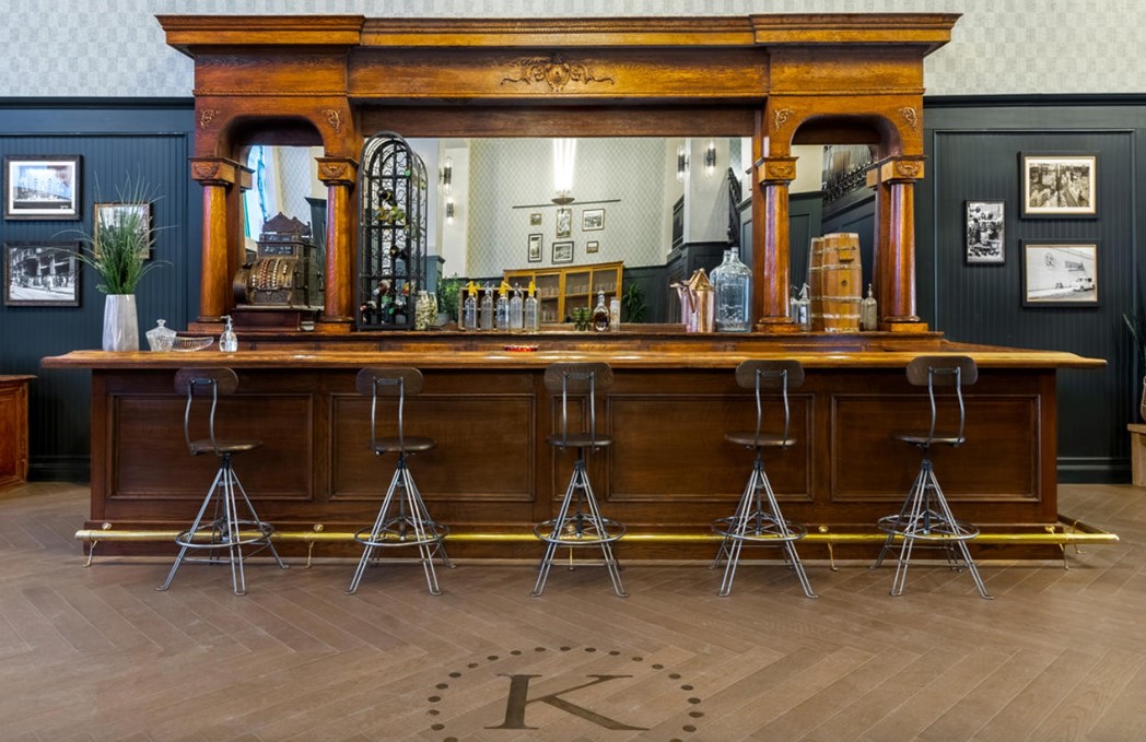 Long wooden bar counter with four metal stools, mirror, and framed pictures on dark walls, wooden floor, and a letter 'K' on the floor.