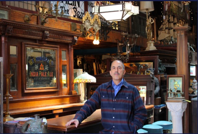Man standing in a bar with wooden furniture and vintage decor, including lamps and framed pictures.