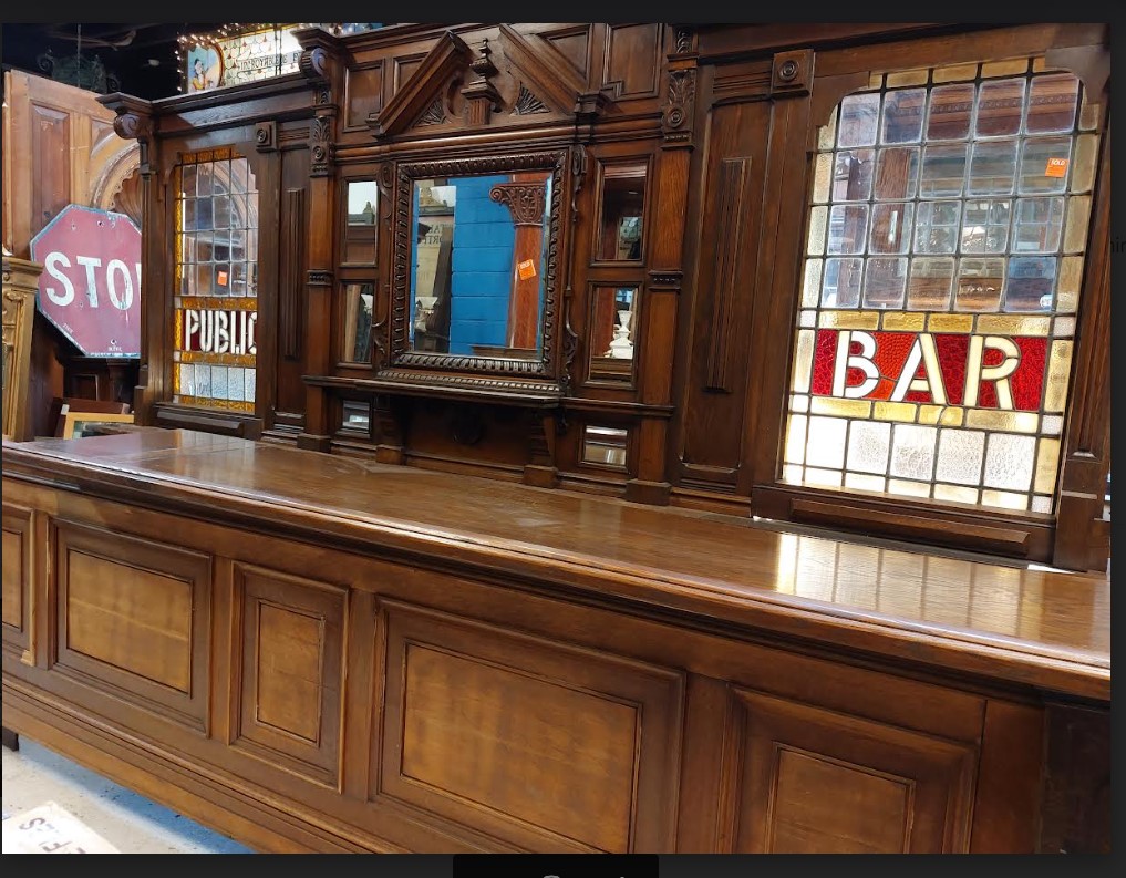 Wooden bar counter with decorative woodwork, mirror, and stained glass window with 'BAR' sign.