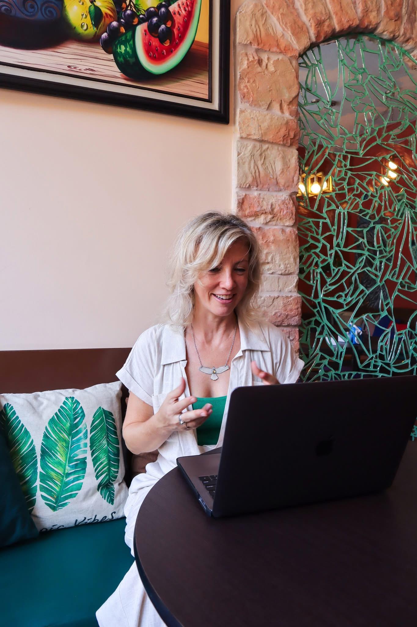 Woman with blonde hair smiling at a laptop, sitting at a round table with a decorative pillow, in a cozy room.