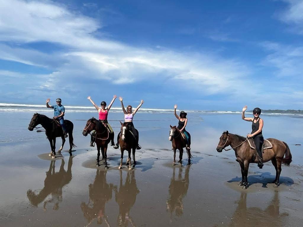 Five people on horseback standing on a beach with water and sky in the background, some with arms raised.