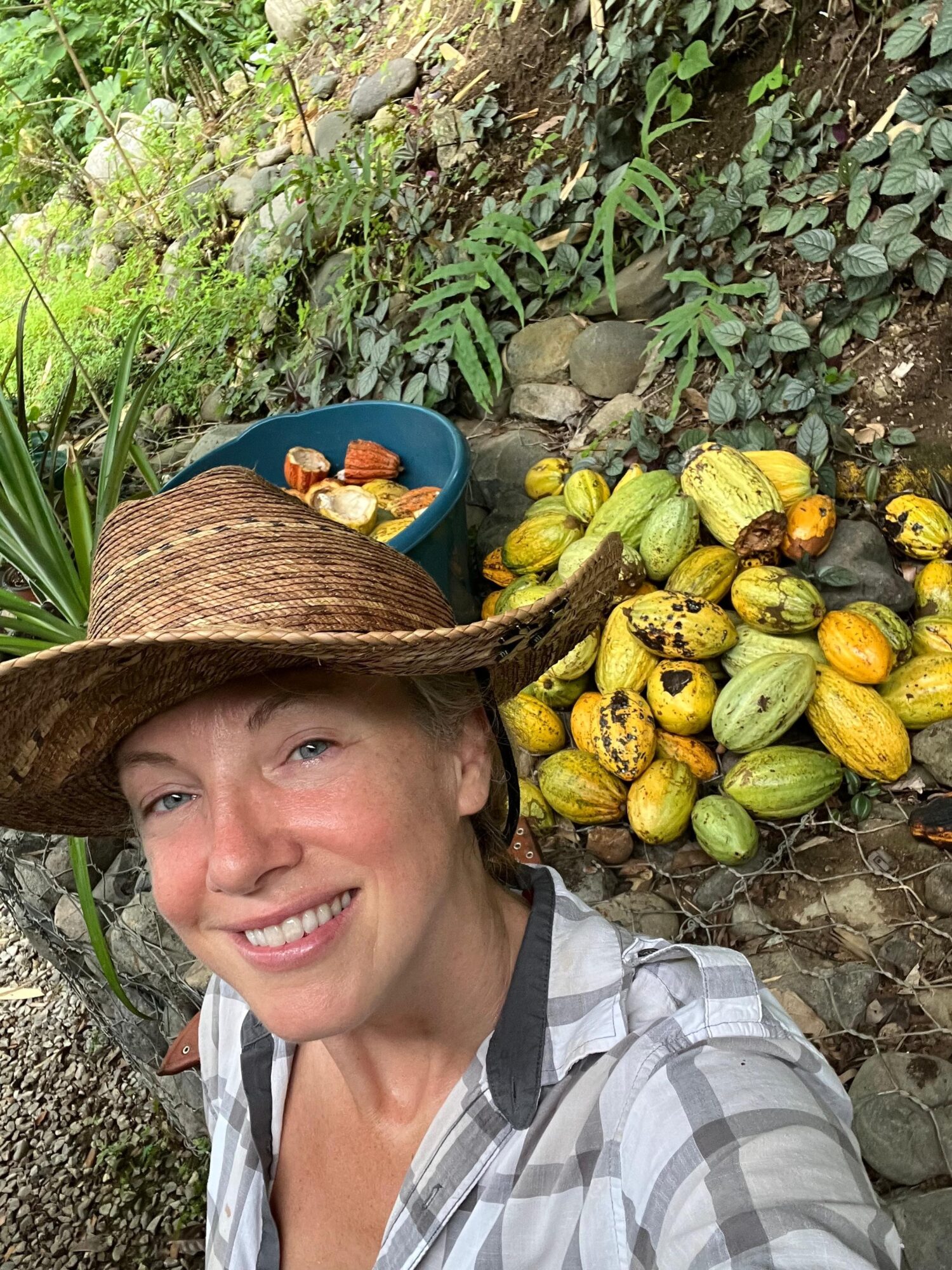 Smiling woman wearing a wide-brimmed hat and plaid shirt in a garden with a pile of yellow and green gourds behind her.