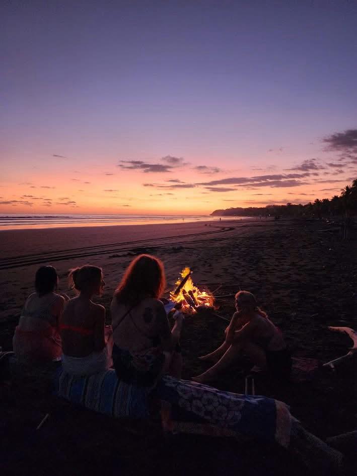 Group of people sitting on a beach at sunset around a small fire, with a colorful sky and ocean in the background.