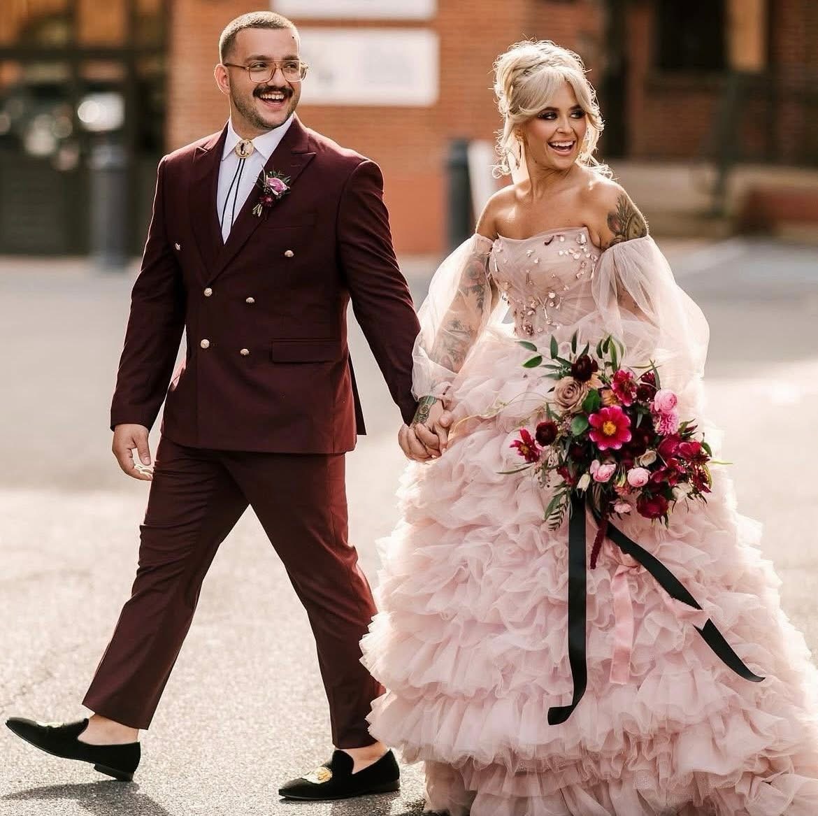 A couple in wedding attire holding hands outdoors, smiling, with a brick building in the background.