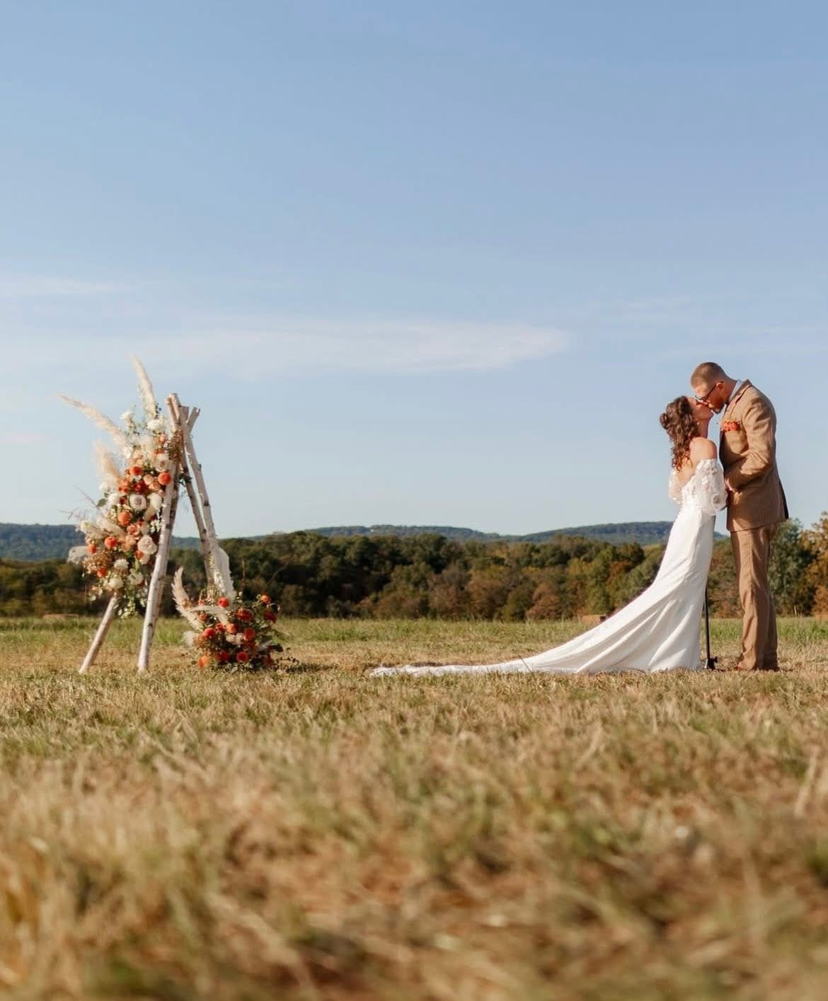 A couple stands outdoors during a wedding ceremony, with a floral arch and open field in the background.