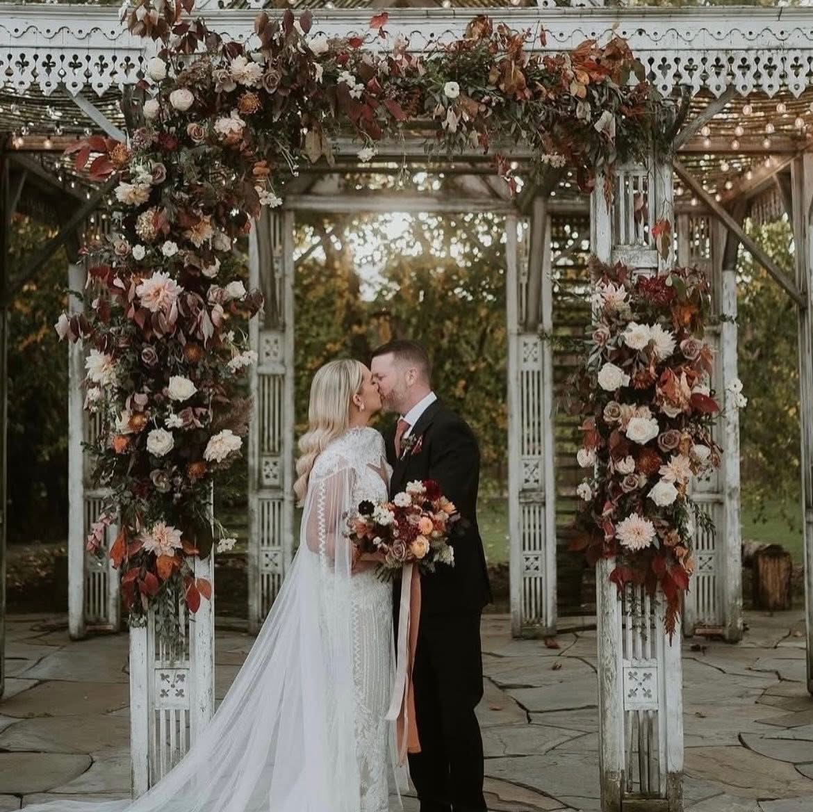 Bride and groom kiss under a floral arch outdoors during sunset, surrounded by trees and a decorative gazebo.