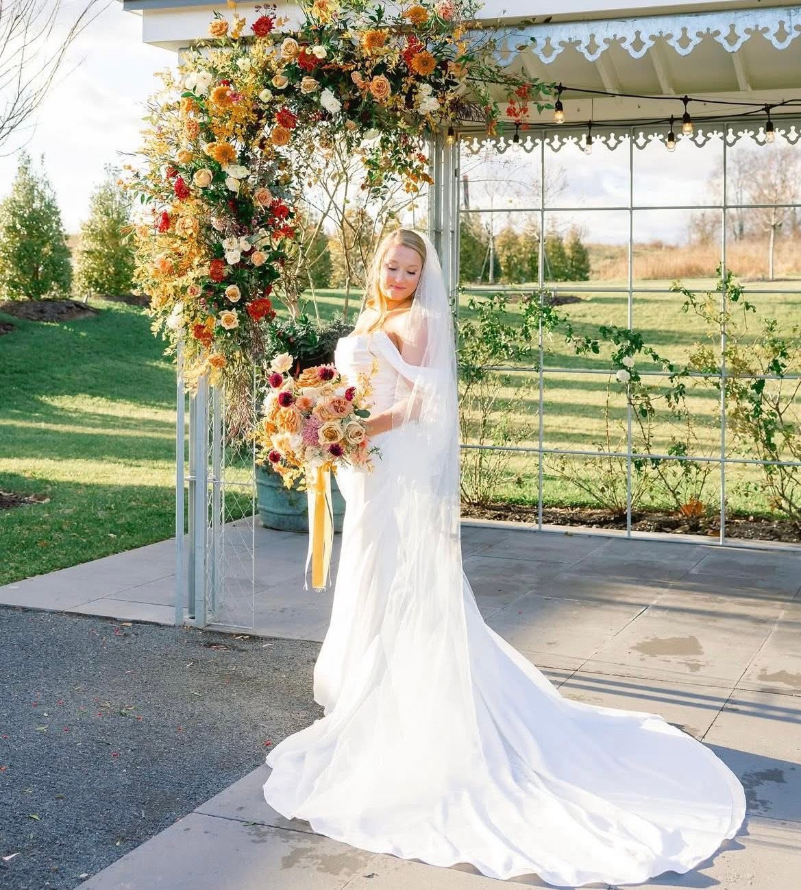 Bride in a white wedding dress holding a bouquet, standing outdoors near a floral arch, with a garden background.