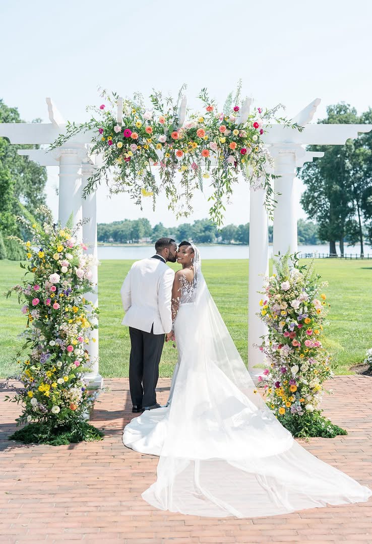 Bride and groom stand under a floral arch outdoors, facing each other, with trees and a field in the background.