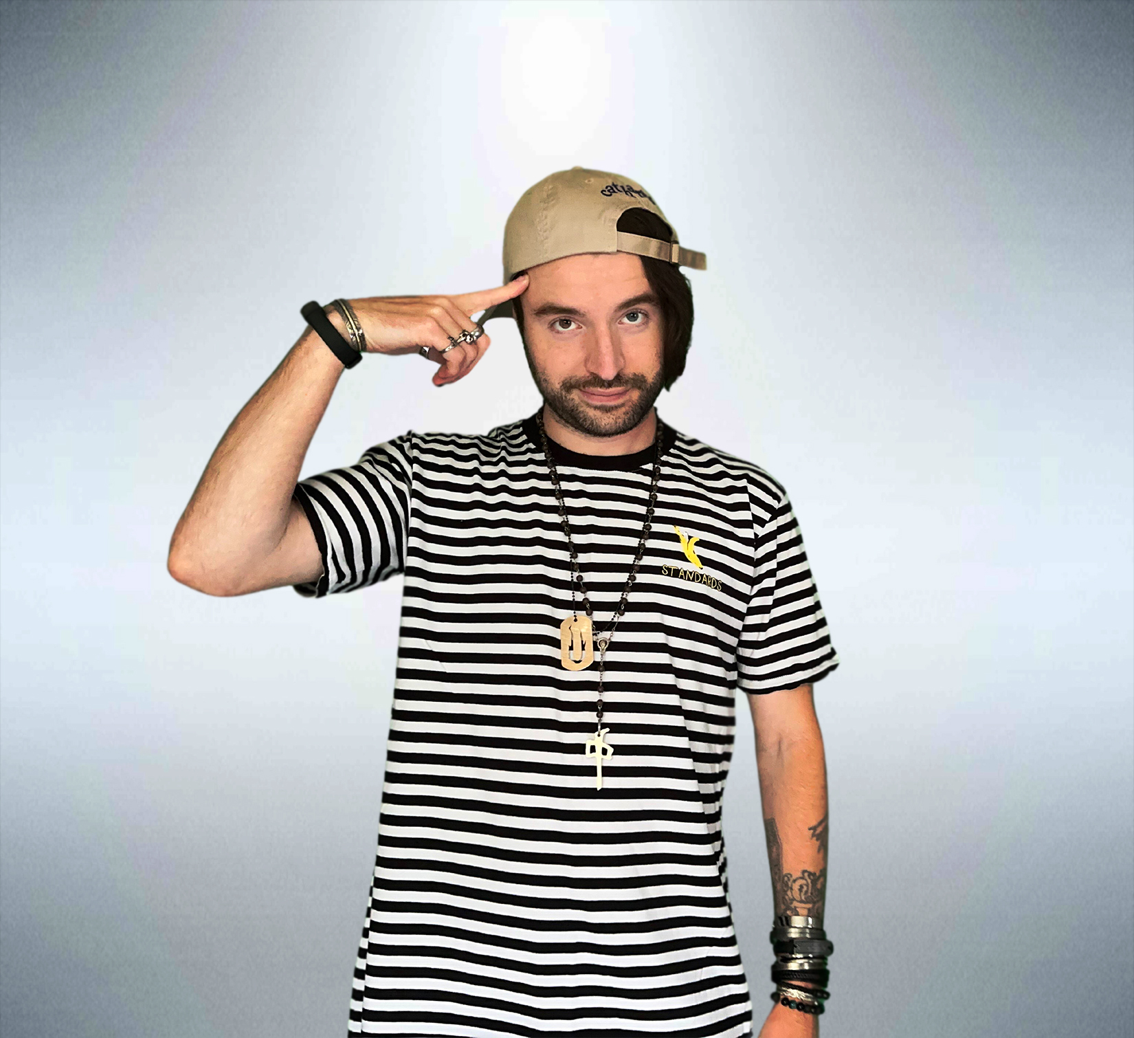 Young man with a beard, wearing a striped t-shirt, beige cap, and accessories, saluting with his right hand.