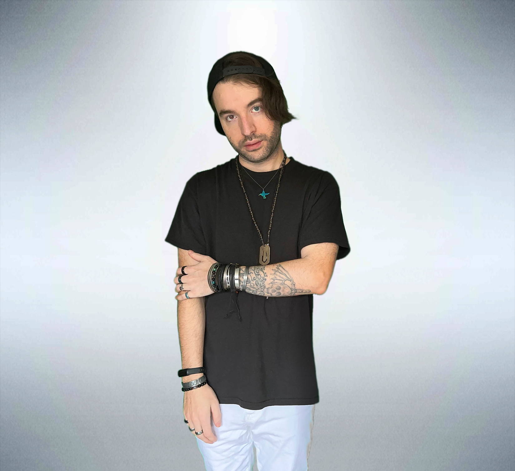 Young man with dark hair, wearing black t-shirt and white pants, standing with arms crossed against a plain background.