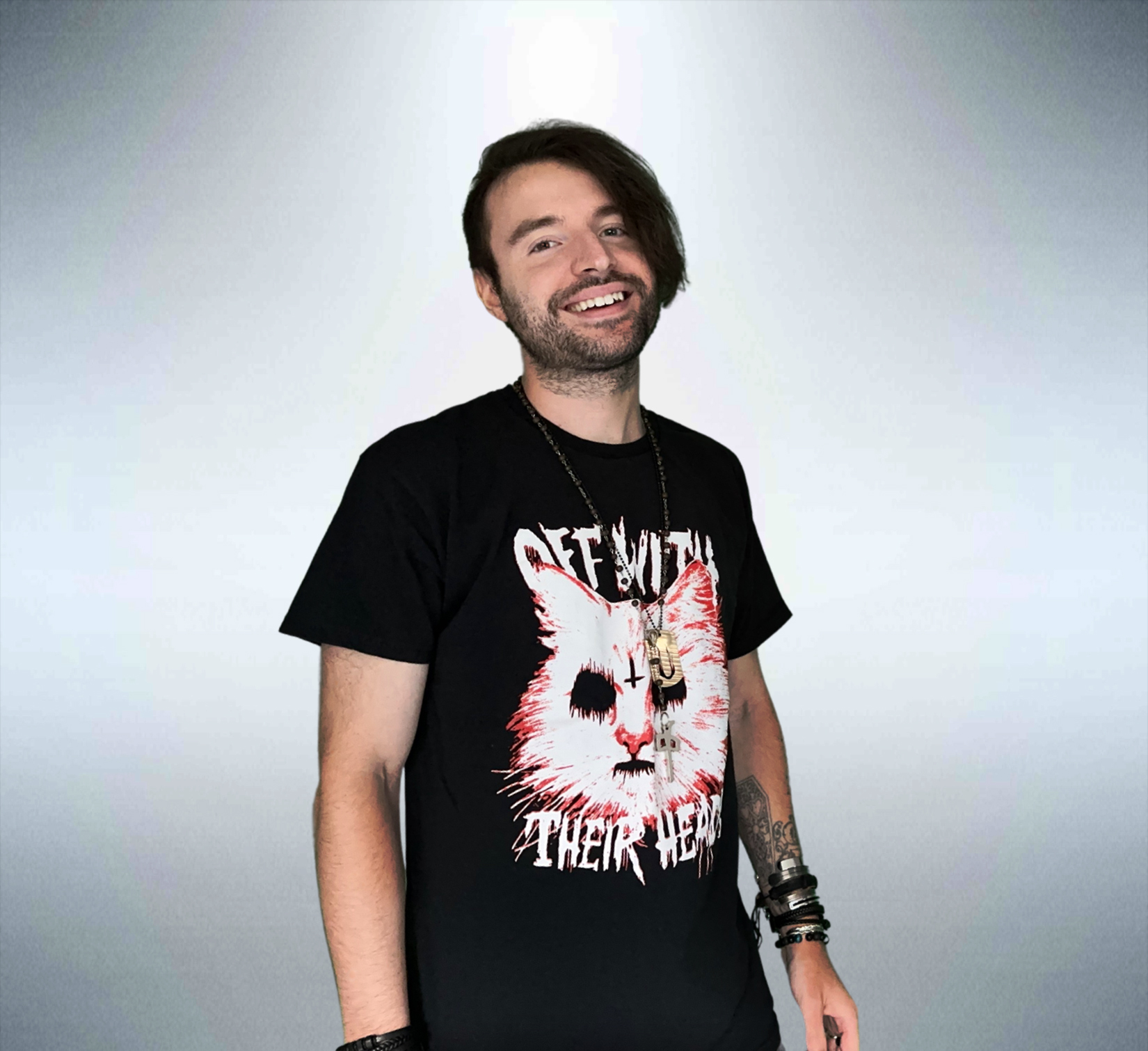 Young man with dark hair and beard smiling, wearing a black T-shirt with a graphic of a cat's face and text, standing against a plain background.