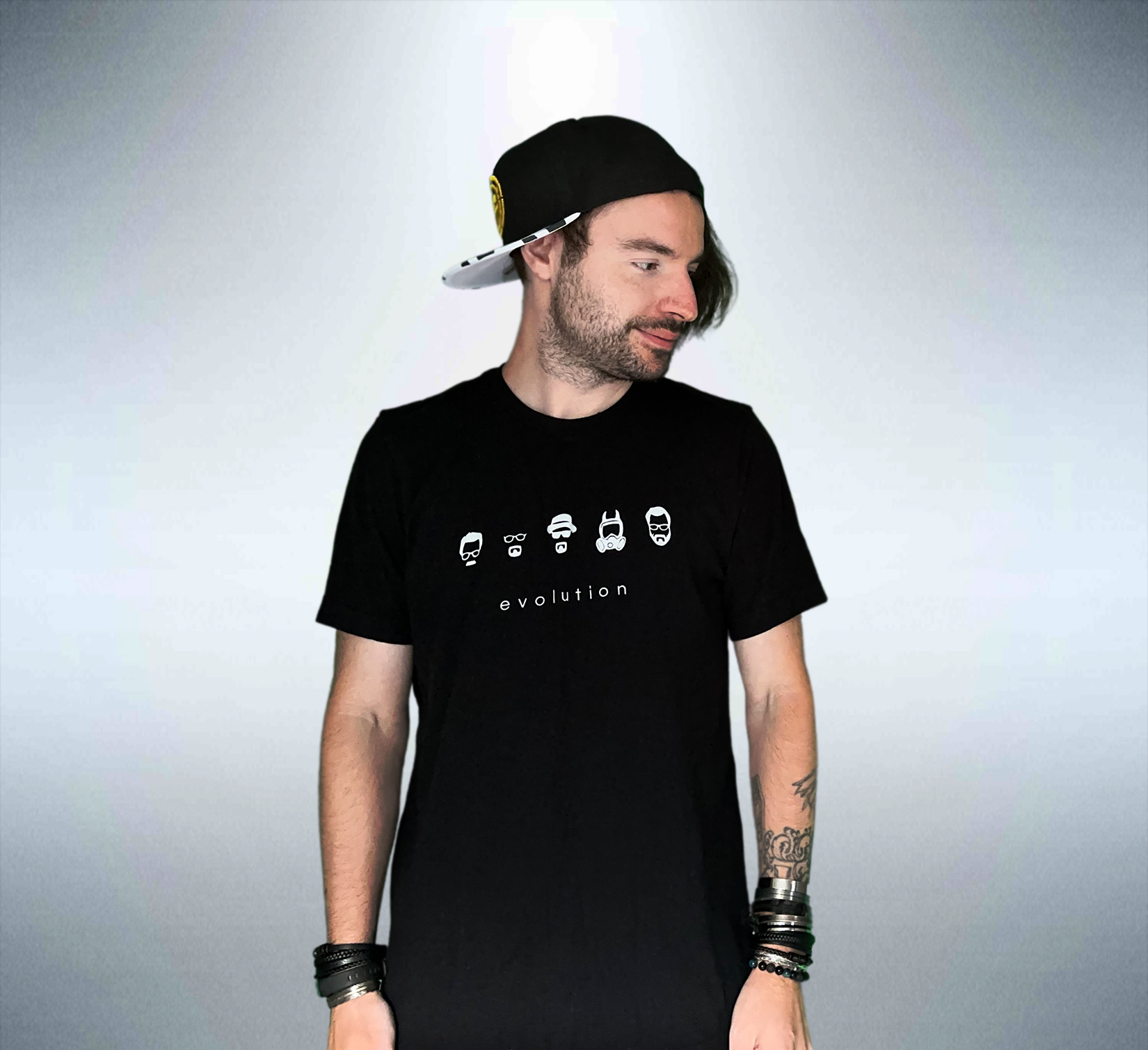 Young man with a beard and tattoos wearing a black t-shirt with car icons and the word 'evolution', black cap, and bracelets, looking to the side.