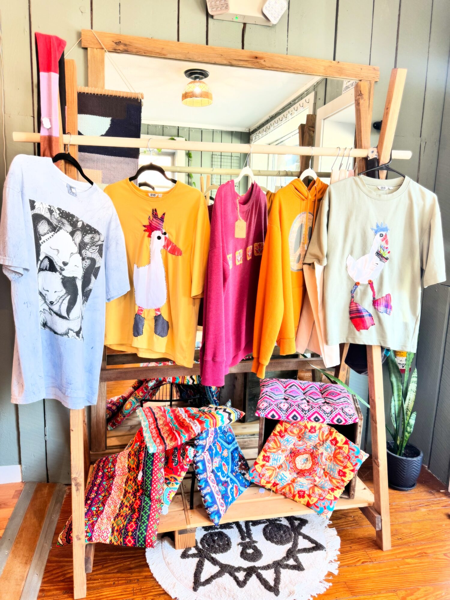 Clothing rack with colorful T-shirts and dresses, pillows with vibrant patterns, and a mirror reflecting the room.