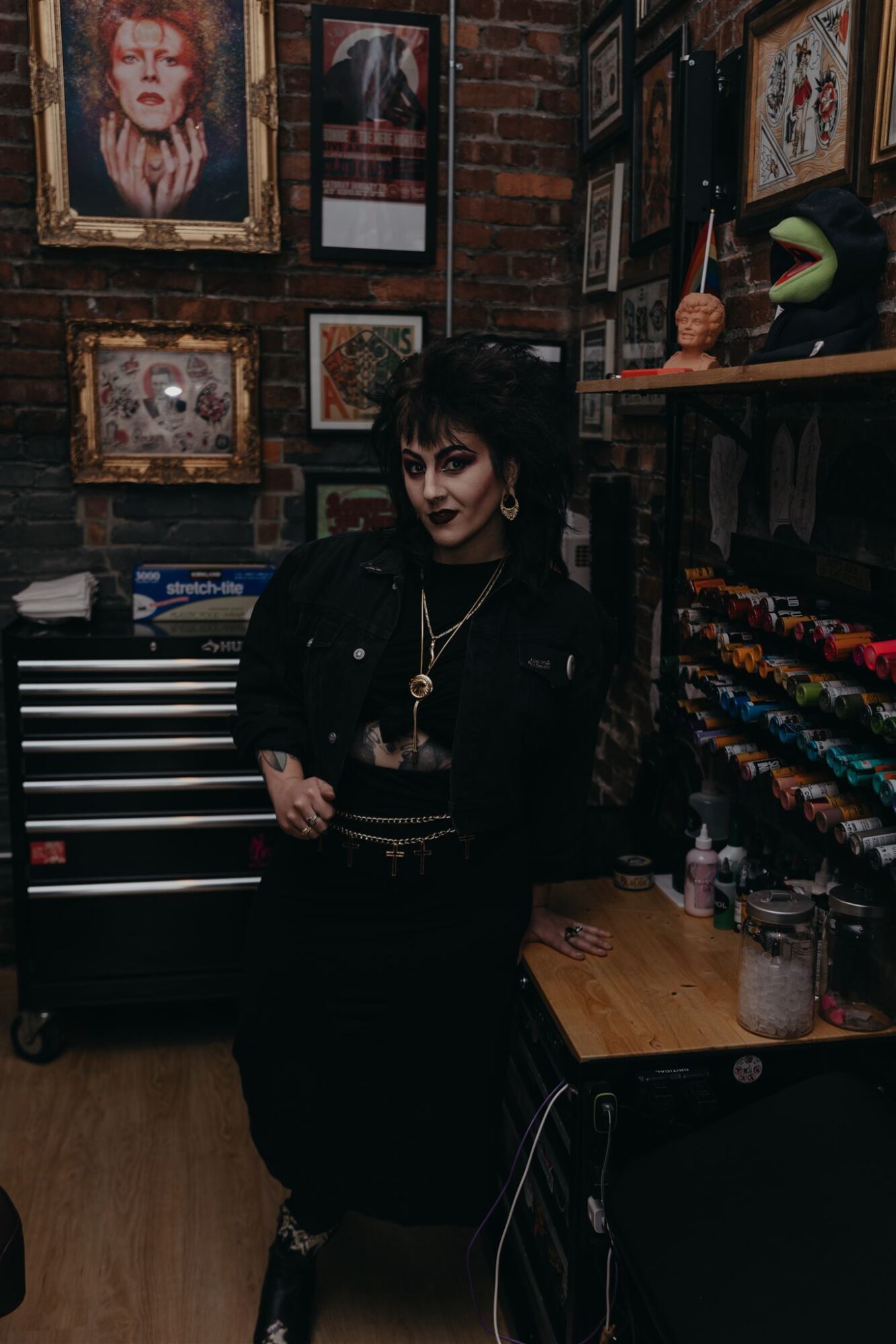 Person with dark hair and makeup standing in art gallery with framed artwork on brick wall behind.