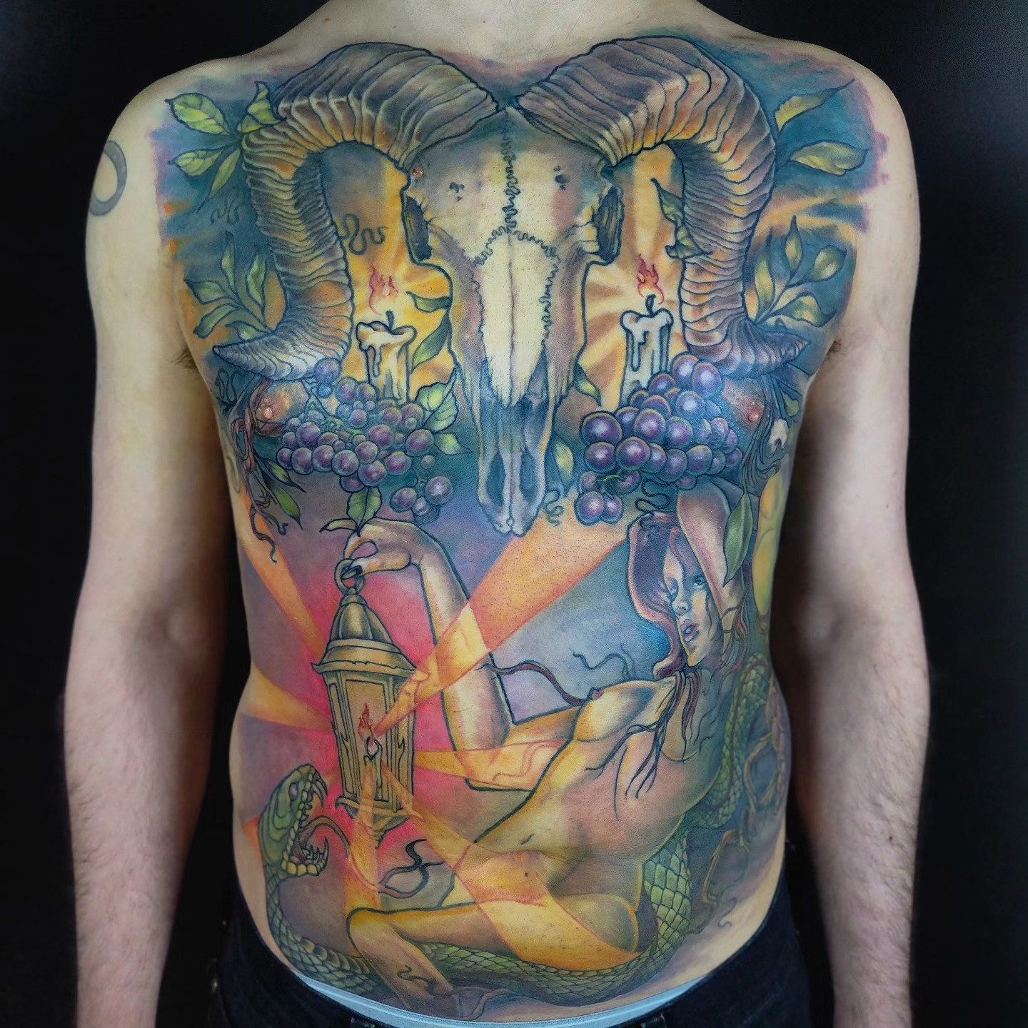 Colorful tattoo of a ram's skull with large curved horns, surrounded by grapes, a lighthouse, and abstract elements on a person's chest and abdomen.
