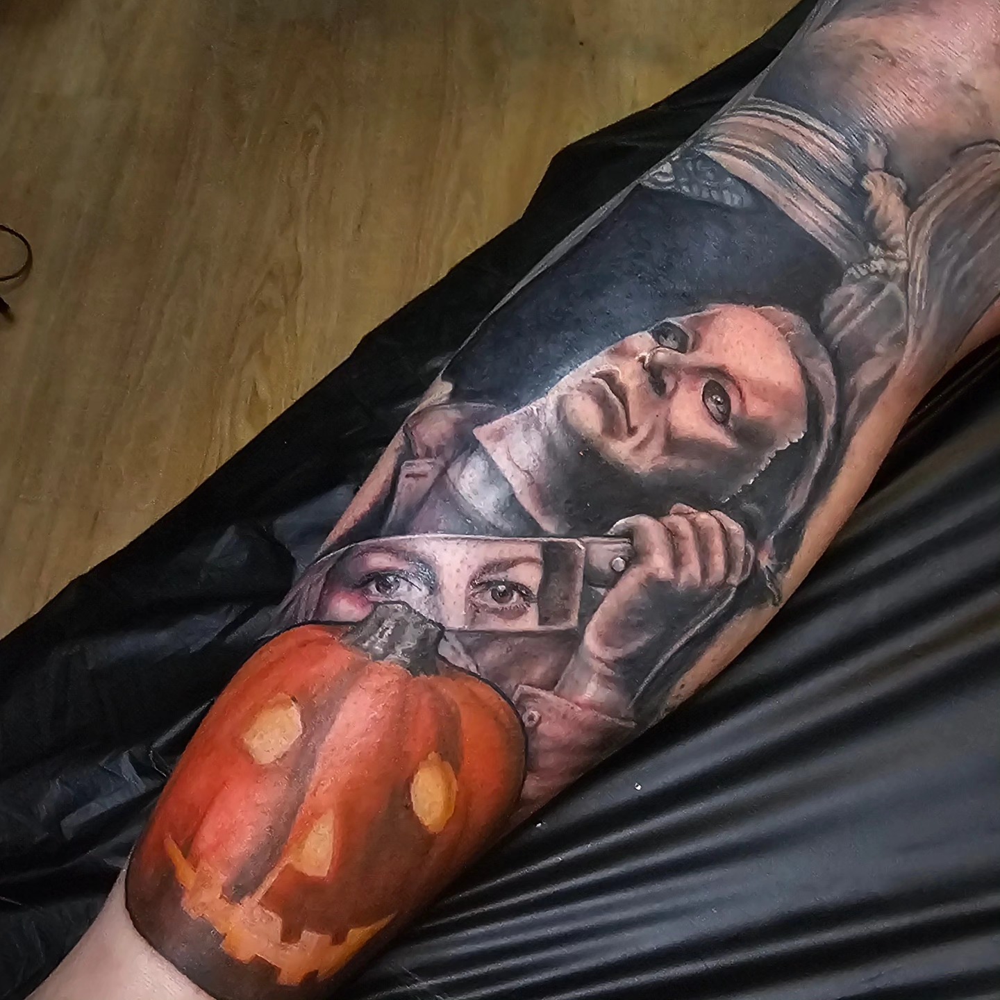 Tattoo of a woman holding a phone with a pumpkin face, with a background of a wooden floor.