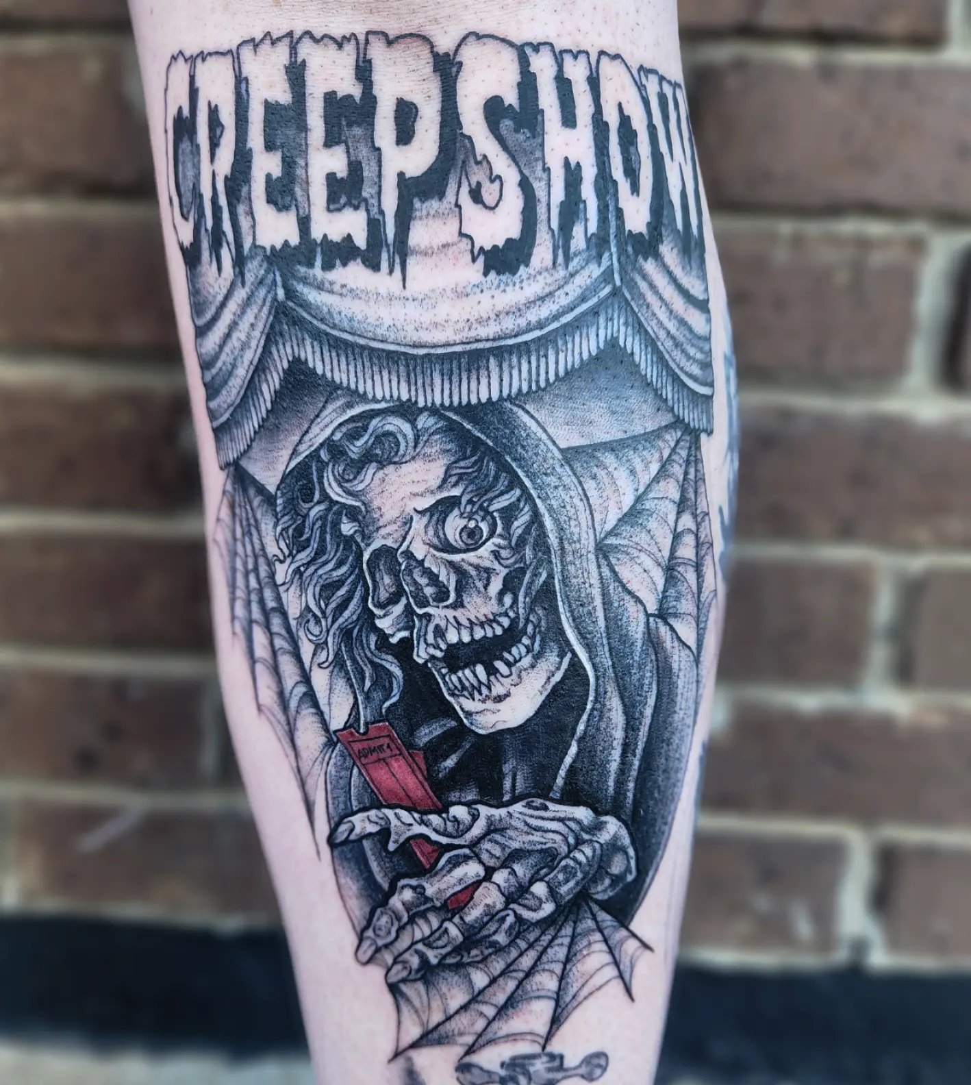 Tattoo of a skeleton with a hood, holding a red object, surrounded by spider webs, with the text 'CREEP SHOW' above.