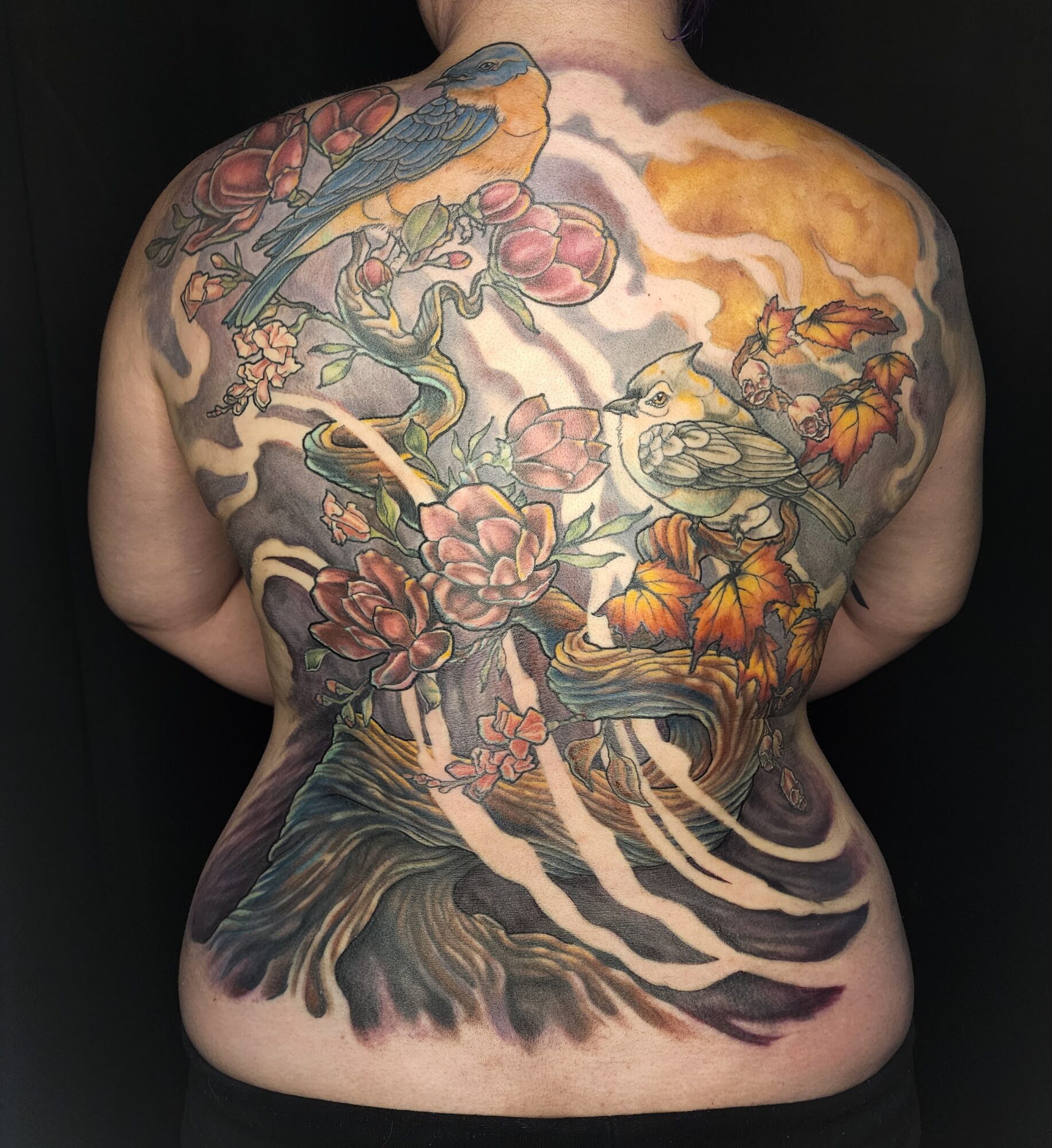 Back tattoo featuring colorful birds, flowers, and a tree with a cloudy sky background.