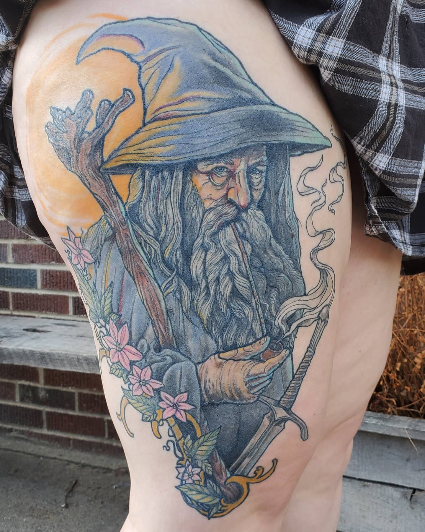 Detailed tattoo of a wizard with a long beard, wearing a pointed hat, holding a staff, surrounded by flowers.
