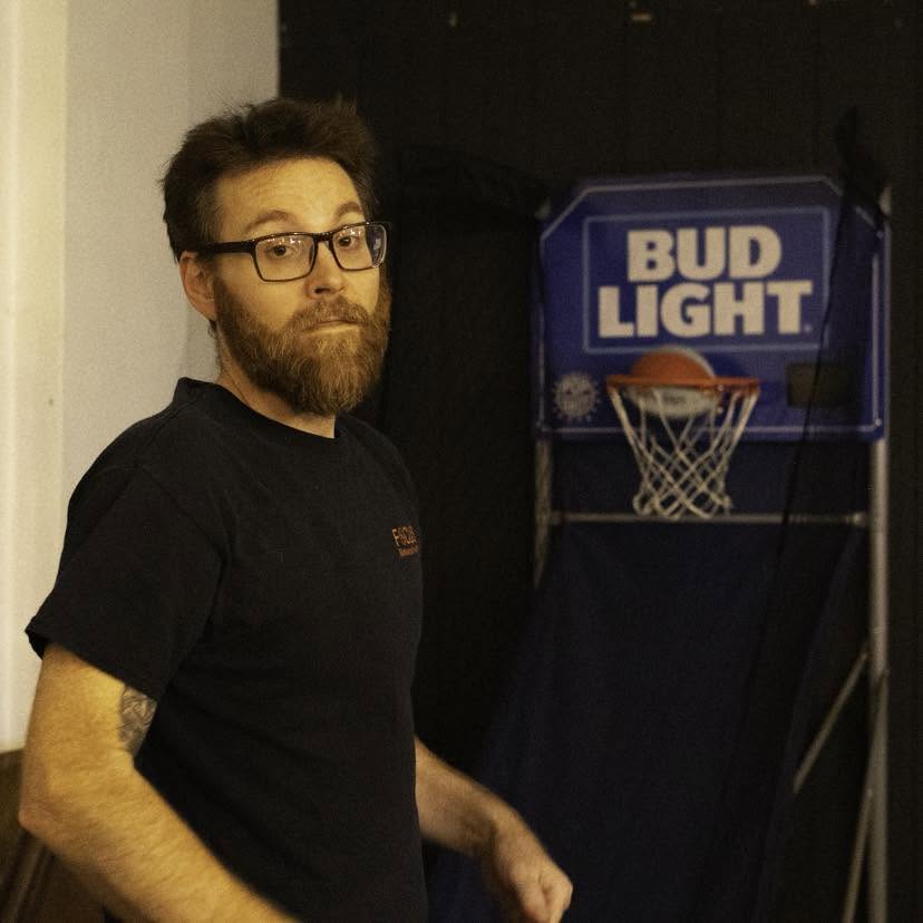 Man with glasses and beard standing near a basketball arcade game, looking at the camera.