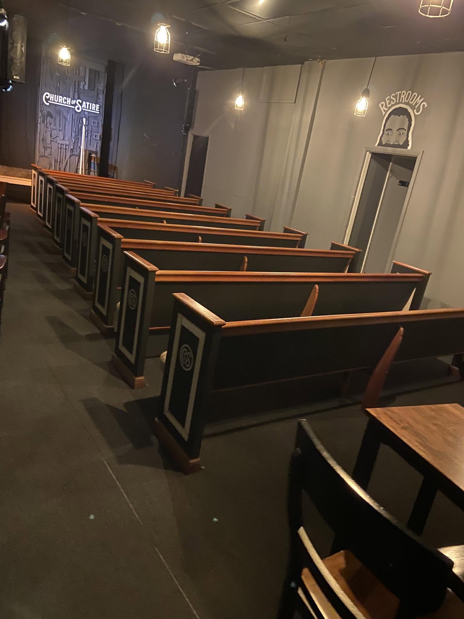 Empty church pews facing a wall with a small arch and a door, dimly lit interior, wooden furniture, dark flooring.